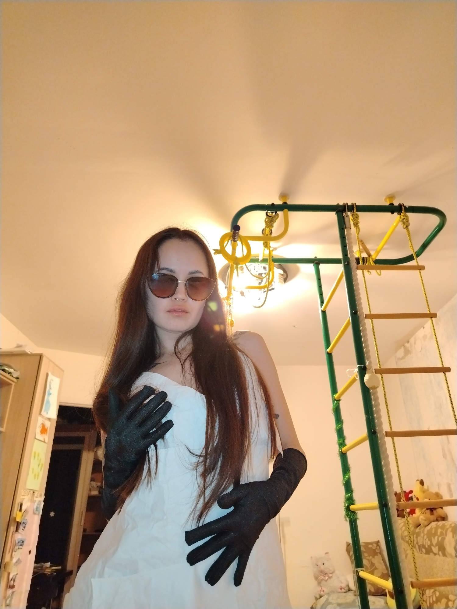 The image captures a woman posing indoors, viewed from a low angle, in what appears to be a domestic setting in Noviy, Russia. The woman has long, dark hair and wears dark sunglasses, a white strapless top or dress, and long black gloves. A nose piercing is visible. She holds her left hand near her chest/shoulder and her right hand slightly extended.
To the woman's right, a prominent green and yellow indoor jungle gym is visible, equipped with yellow rings, horizontal bars, and a rope ladder with wooden rungs. Sections of the jungle gym are decorated with green tinsel. Above, a multi-bulb ceiling light fixture illuminates the room, casting light onto the light-colored ceiling.
In the background on the left, a wooden cabinet or shelving unit is present, adorned with several pinned drawings or papers. On the right side of the background, behind the jungle gym, there is furniture, possibly a sofa or bed, featuring pillows and several stuffed animals, including a Pikachu and a white cat. The presence of the jungle gym and toys suggests a child's room or family play area. Faint, illegible Cyrillic text is visible on the cabinet on the left. The lighting in the scene is provided by internal artificial sources.