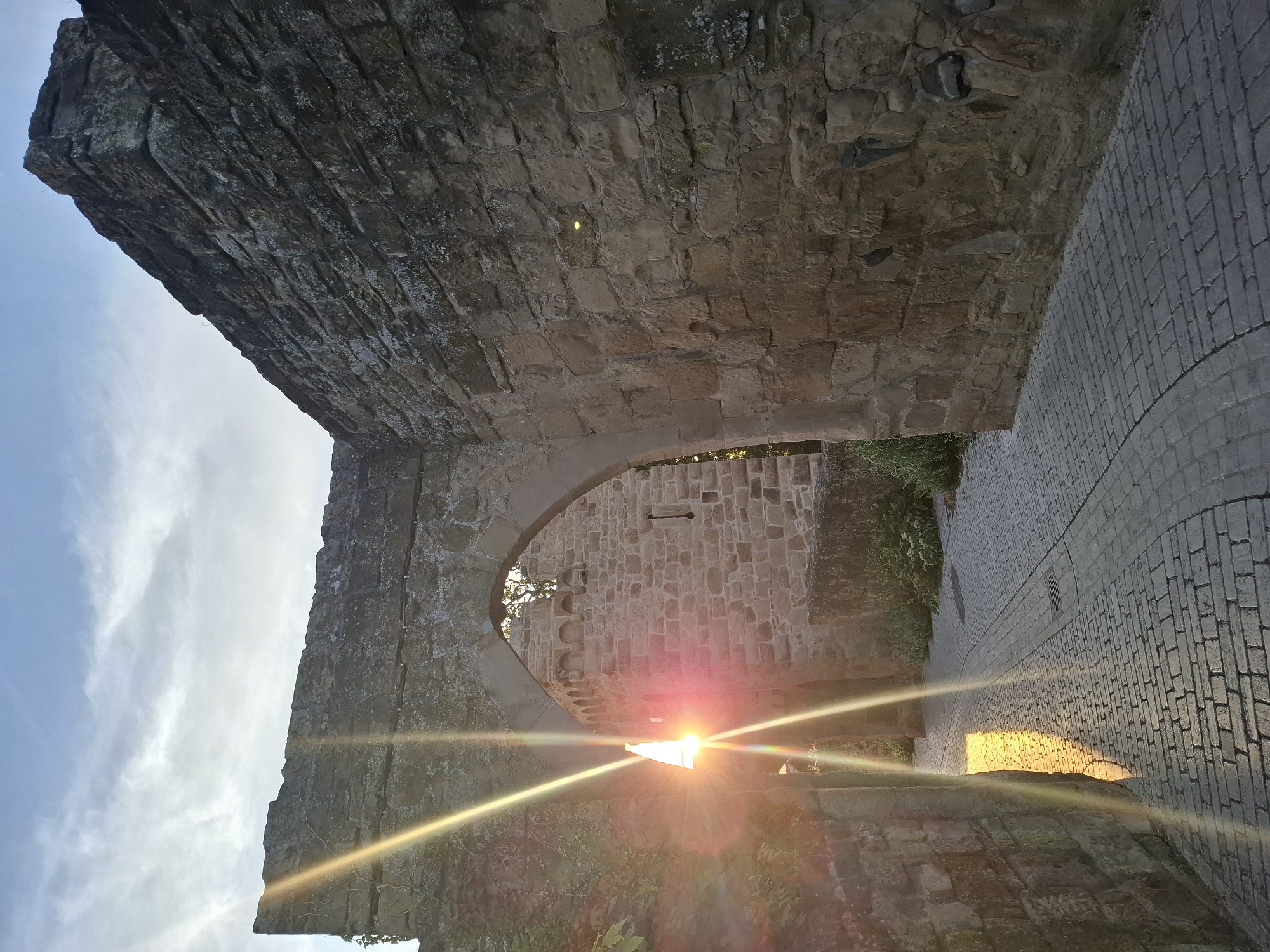 The photograph captures a sun-drenched stone archway within the historic city walls of Sinsheim, Germany. The setting sun casts a warm, golden glow, creating dramatic light rays that streak across the scene. The stone walls, likely part of an ancient fortress or gate, are constructed from rough-hewn blocks, showing signs of age and weathering.
Through the arch, another stone structure is visible, possibly a tower or inner courtyard, also built with weathered stone. The ground is paved with intricate brickwork, forming a pathway that leads through the archway. Lush green vegetation grows along the base of the walls, adding a touch of nature to the ancient architecture. The sky above is a mix of pale blue and wispy white clouds, illuminated by the setting sun. No people are present in the image. The overall atmosphere is one of historical grandeur and serene beauty.