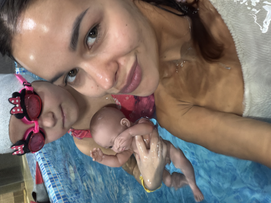 The image displays an adult female and a child in an indoor swimming pool. The specific location within Krasnoyarsk, Russia, cannot be determined from the visual information.
The adult female, positioned on the right, faces the camera directly. She has dark hair, brown eyes, and tan skin, and appears to be submerged in the pool up to her chest, with water visible around her shoulders. Her hair is wet, and she is wearing a white towel or garment partially submerged in the water.
To her left and slightly behind her, a child is visible, also in the water. The child wears a white swimming cap and pink swim goggles adorned with black and red Minnie Mouse ear designs. The child is wearing a pink swimsuit top. The child is holding a light-skinned baby doll, which is partially submerged and appears to be wearing yellow swim bottoms; the doll's arms are bent upwards towards its face.
The swimming pool water is clear and blue with visible ripples. A tiled pool edge, with blue and white patterned tiles, is visible in the background on the left. A pair of red swim fins is partially visible on the far left edge of the pool.