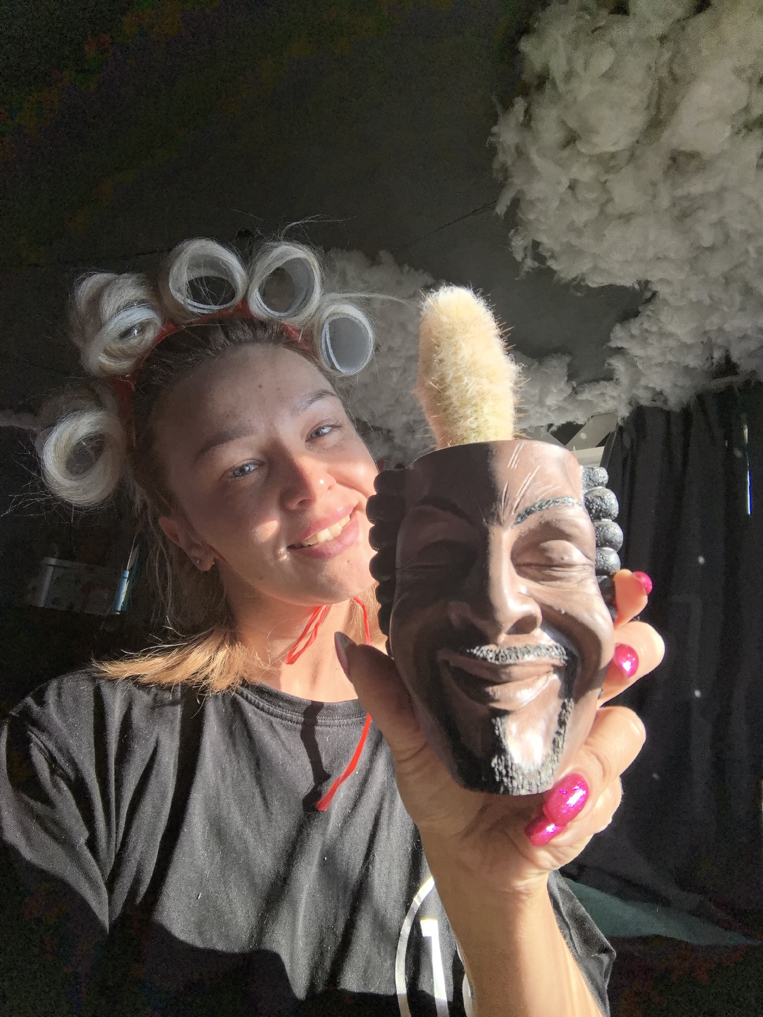 A young woman is holding up a pot shaped like the face of Snoop Dogg with a cactus planted inside. She is smiling and wearing hair rollers. The scene appears to be inside, with what looks like artificial clouds overhead. I cannot confirm the location in the city of Noviy, Russia, without additional information.