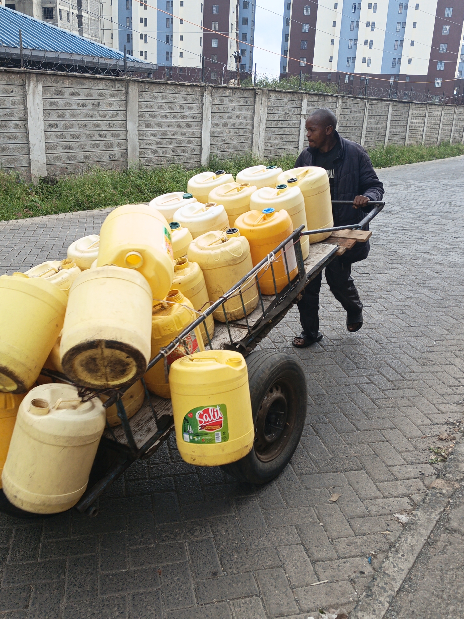 A man pulls a heavily loaded cart filled with yellow plastic containers along a brick-paved road in Nairobi, Kenya. The containers appear to be full of liquid, possibly water. The cart has one visible wheel. The man is wearing dark clothing and sandals. Behind him is a tall, light-colored building with a blue roof, topped with barbed wire. A concrete block wall also topped with barbed wire runs along the side of the road. The sky is overcast.