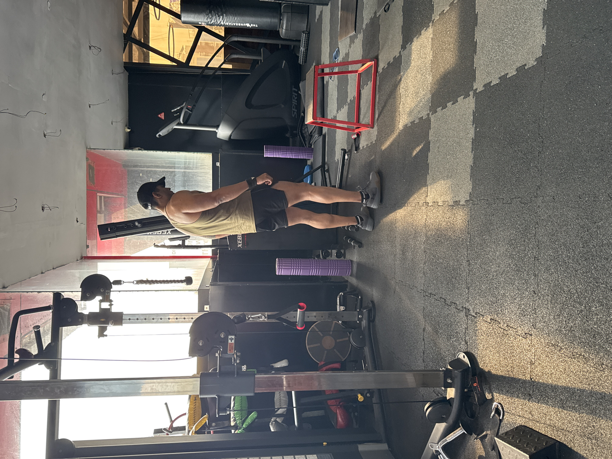 The image shows a man standing in a gym in Kolkata, India. He is wearing a tank top, shorts, and a baseball cap. He is standing next to a weight machine, and there is a treadmill and a plyometric box in the background. The gym has gray and black interlocking floor tiles. The walls are white and there are some wires running along them. There is also a window with a metal frame.