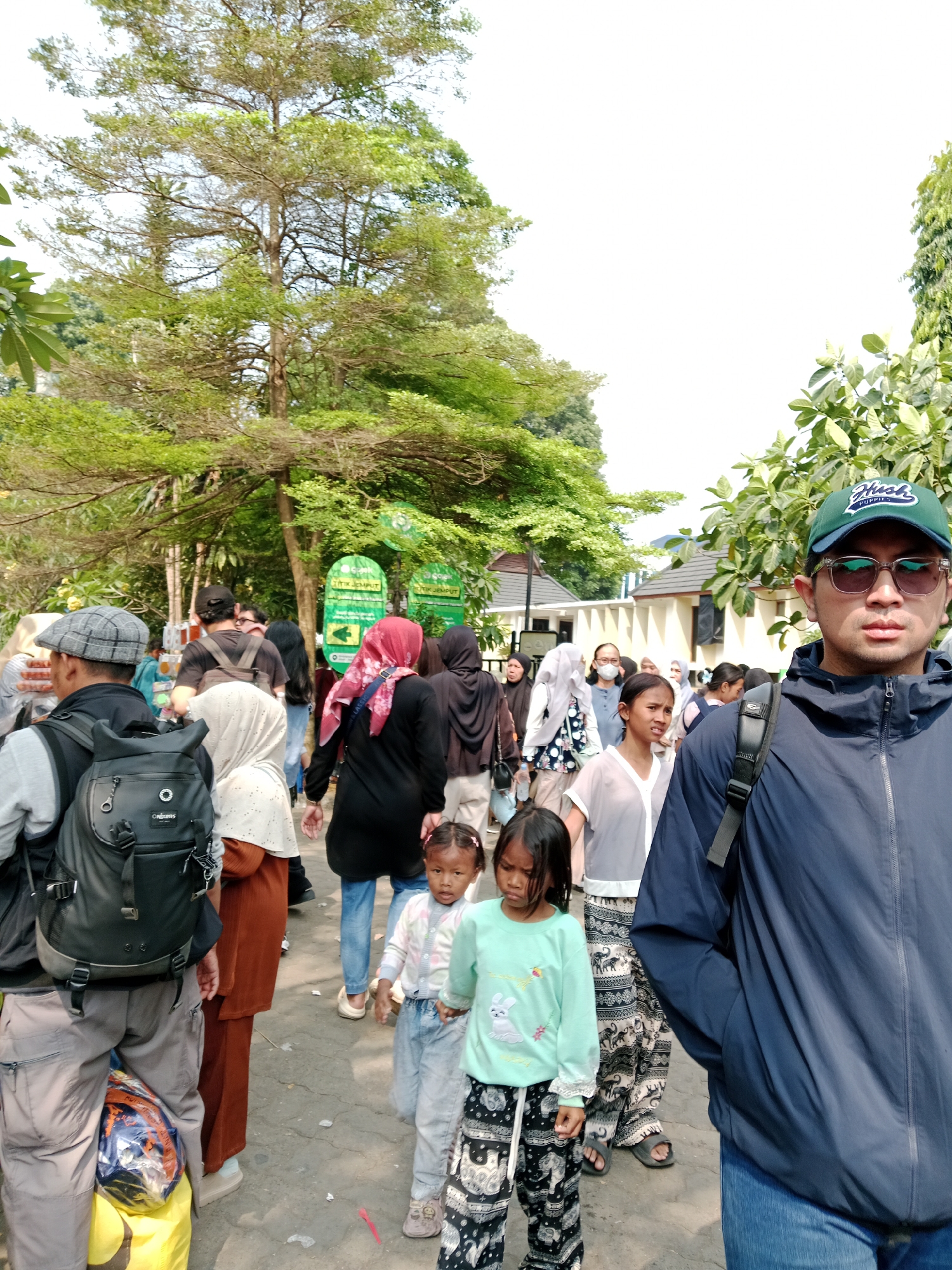 The image depicts a street scene in Bogor, Indonesia, with a crowd of people walking along a paved pathway. In the foreground, a man wearing a dark blue jacket, a green baseball cap, and sunglasses is taking a selfie. He is positioned slightly to the right of the frame, with a large portion of his body visible. Behind him, a group of people are walking in the same direction. Among them are several women wearing headscarves, children, and men.
The environment is characterized by lush greenery, with large trees lining the pathway. In the background, there are some low-rise buildings. Green signs with text are visible on the left side of the pathway, indicating a public area. The lighting suggests it is daytime, likely under bright sunlight, as indicated by the shadows and the washed-out sky. The overall impression is that of a busy public gathering or a tourist area.