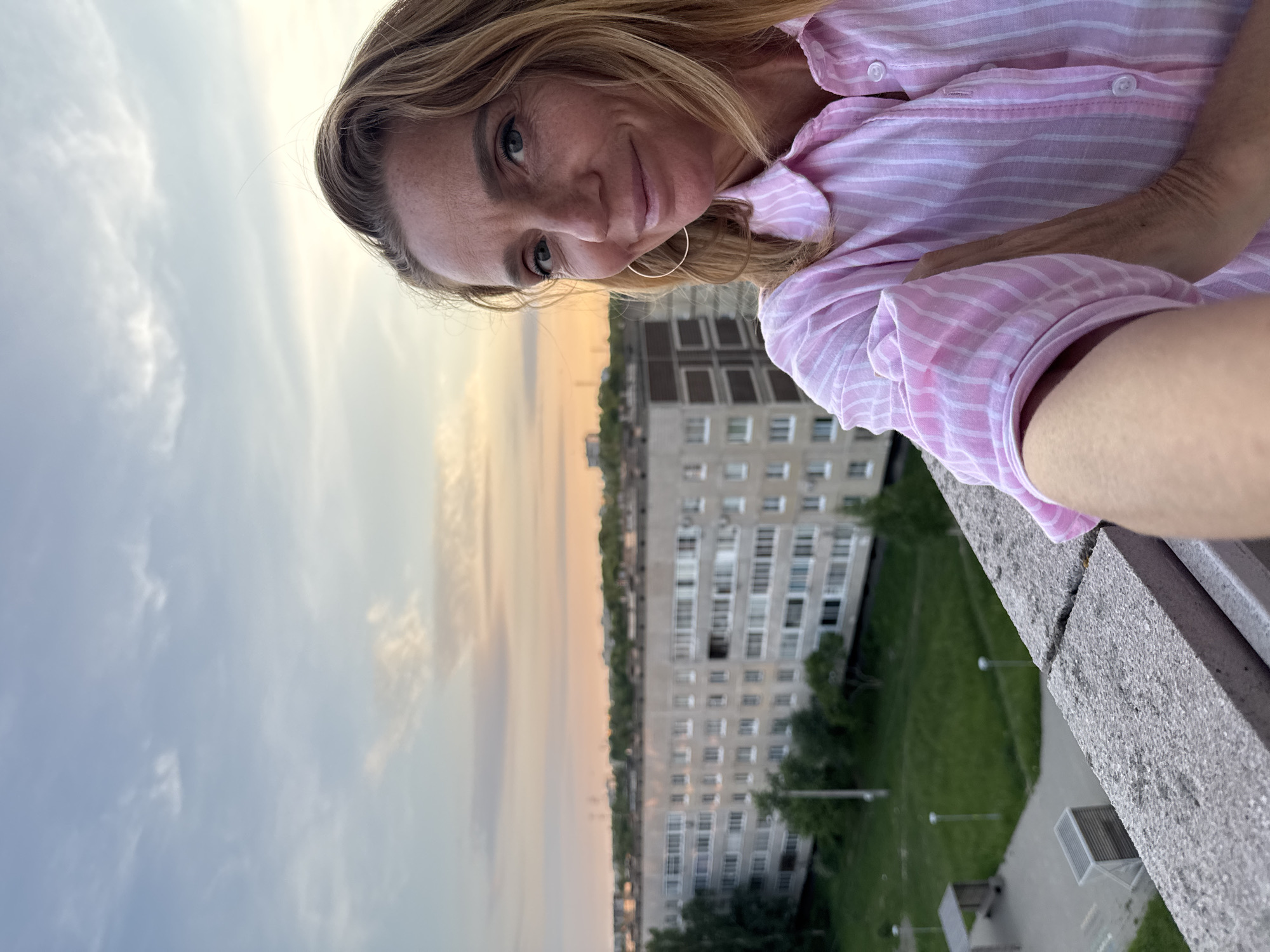A woman with blonde hair and a pink and white striped shirt takes a selfie outdoors in Kirishi, Russia. She is standing on a concrete balcony overlooking a residential area with a multi-story building and green spaces. The sky is a pale blue with streaks of pink and orange, suggesting it is either sunrise or sunset. The woman is smiling and looking at the camera.