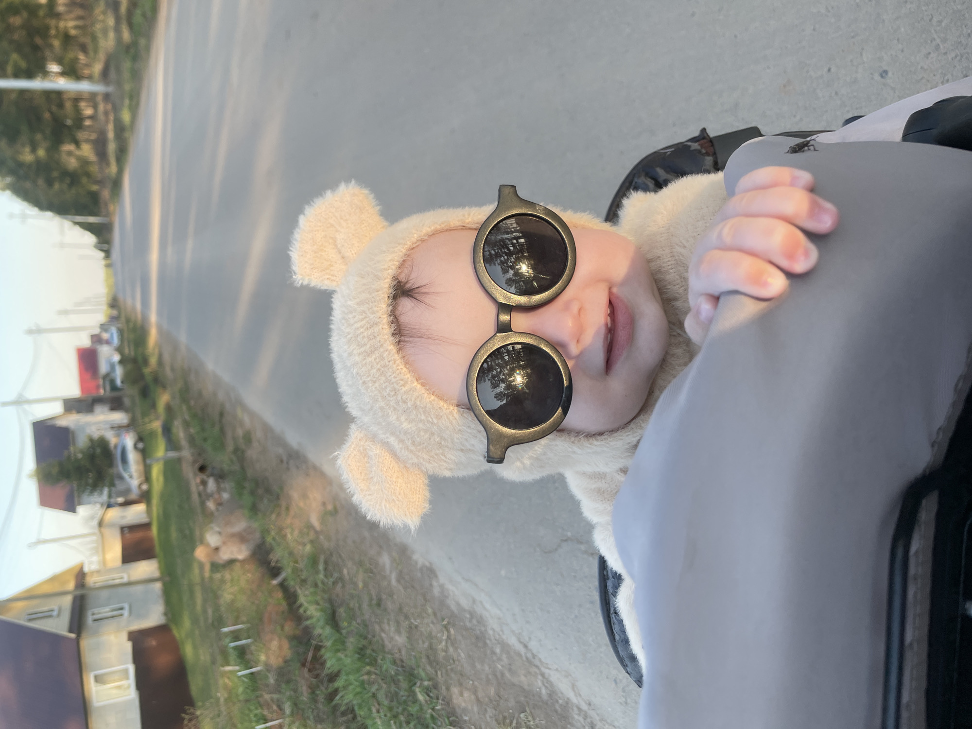 A close-up shot, taken at an unusual angle, shows a baby wearing a fluffy bear-eared hat and dark sunglasses, sitting in what appears to be a stroller. The location appears to be in Подшивалово, Russia. Houses and trees are visible in the blurred background, suggesting a residential area. A gray stroller handle and some of the stroller are visible on the right side of the frame. A dog sits on the side of the road.