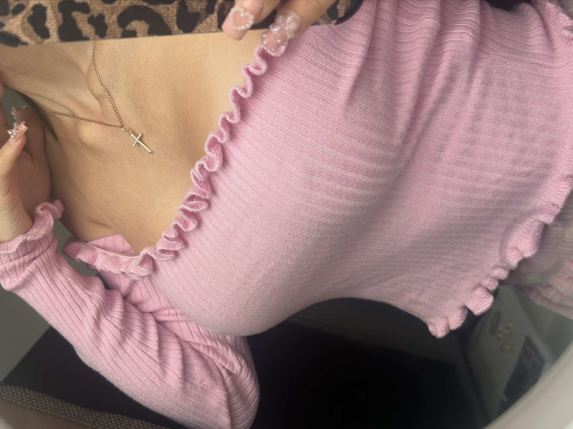 The image shows a close-up view of a person wearing a pink, ribbed, off-the-shoulder top with ruffled edges. A gold cross necklace with small stones is visible around their neck. The person's hand, adorned with sparkly, embellished nails, is visible near their face and the neckline of the shirt. The background is dark and out of focus. There is no visible indication of the city of Rezvykh, Russia, or any elements that could suggest a specific urban location.