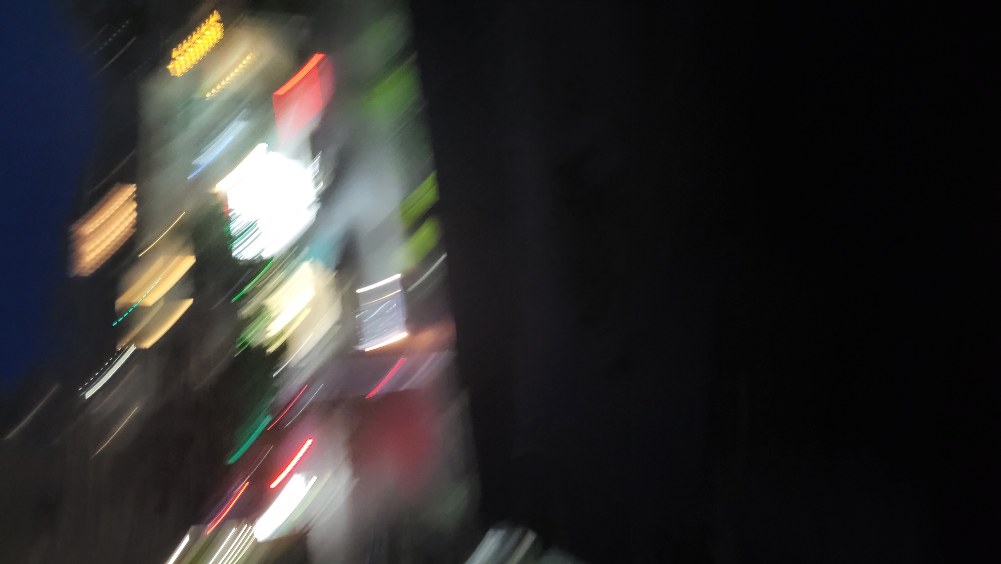 The image depicts a nighttime urban scene, heavily blurred by motion or long exposure, observed from an elevated vantage point. The lower half of the frame is dominated by an indistinct, dark surface, likely a rooftop or balcony railing, which forms a horizontal dark boundary across the mid-section. Above this boundary, the upper half of the image displays a vibrant array of streaked and blurred lights against a dark blue twilight or night sky.
In the upper-middle section, a large, intensely bright white light source, somewhat circular in form, is prominent, flanked by and intermingled with distinct green light streaks. To the left of this central illumination, multiple diagonal streaks of vivid white and red lights are visible, indicative of moving vehicle headlights/taillights or streetlights. Towards the upper-right, several parallel, diagonal streaks of warm yellow-orange light suggest illuminated building facades or specific commercial signs. Due to the significant motion blur, no specific subjects, objects, or people are clearly discernible. The overall impression is that of a bustling commercial area at night.
Based on the visual characteristics, particularly the prominent white and green light configuration resembling a known coffee shop chain's signage, and the general urban nightscape, the scene is likely located in the city of Yerevan, Armenia. The specific viewpoint appears to be overlooking a busy commercial street such as Northern Avenue, from an elevated position, capturing the distinctive lights of establishments like "The Coffee Bean & Tea Leaf" and surrounding traffic.