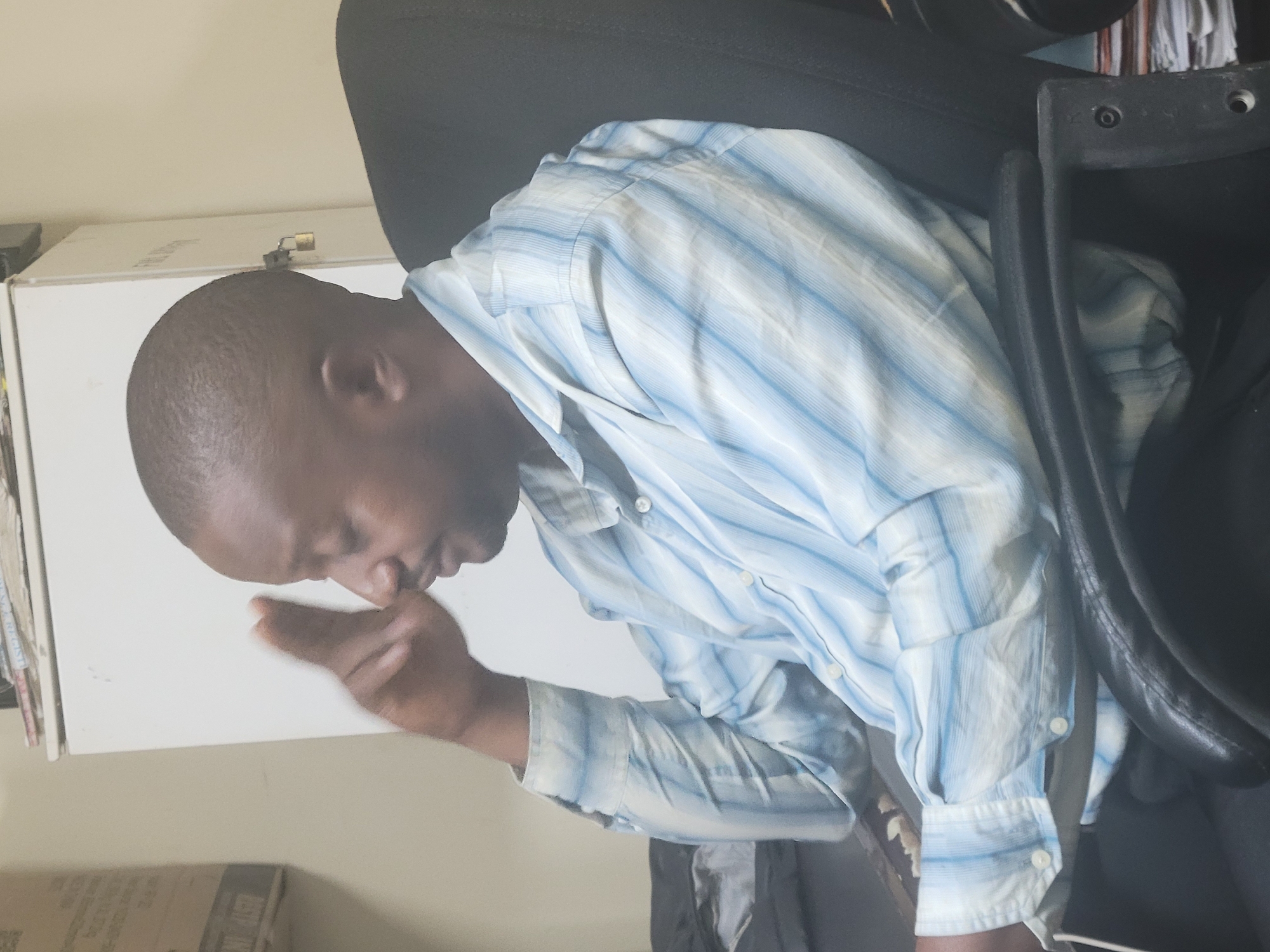 In a dimly lit room, likely an office in Abuja, Nigeria, a man sits in a black office chair. He is wearing a light blue and white striped button-down shirt. His right hand is raised to his temple, with his index finger extended, suggesting he is in thought or experiencing a headache. His gaze is directed downwards and to the right. Behind him, a white cabinet with a small padlock is visible. To the left, a cardboard box is partially seen, and a stack of papers is visible on a shelf above. The overall atmosphere is one of quiet contemplation or a moment of pause.