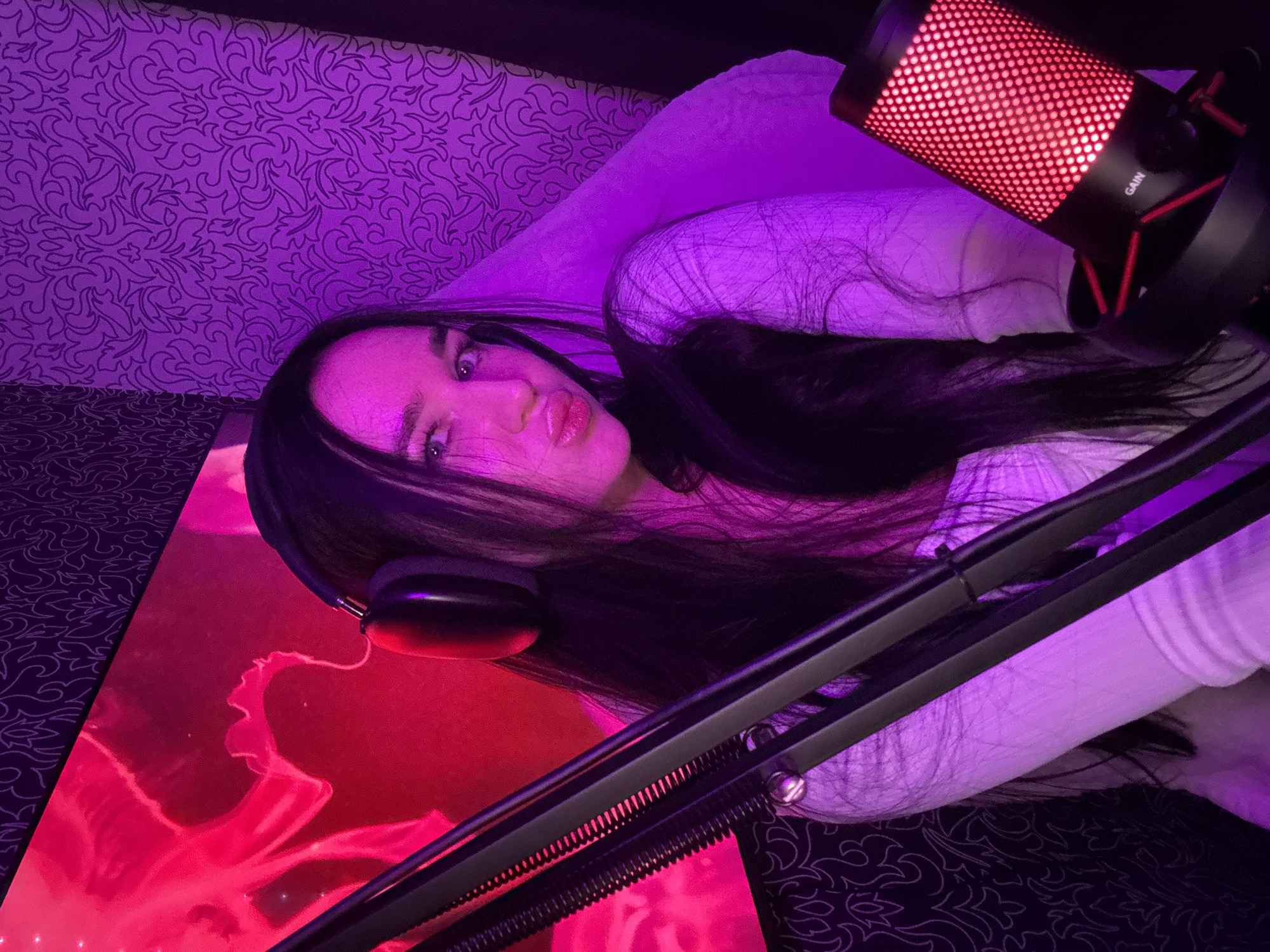 A young woman with long dark hair and headphones is posing for a picture in a dimly lit room. She is wearing a light-colored top and is looking directly at the camera with her lips pursed. To her left is a microphone stand, and to her right, a microphone with a red illuminated grill. Behind her, a screen displays a vibrant pink image, possibly of a jellyfish, and the wall is covered in patterned wallpaper. The room is illuminated by pink and purple lighting, creating a dramatic effect. There is no visible indication of the location being in Novosibirsk, Russia.