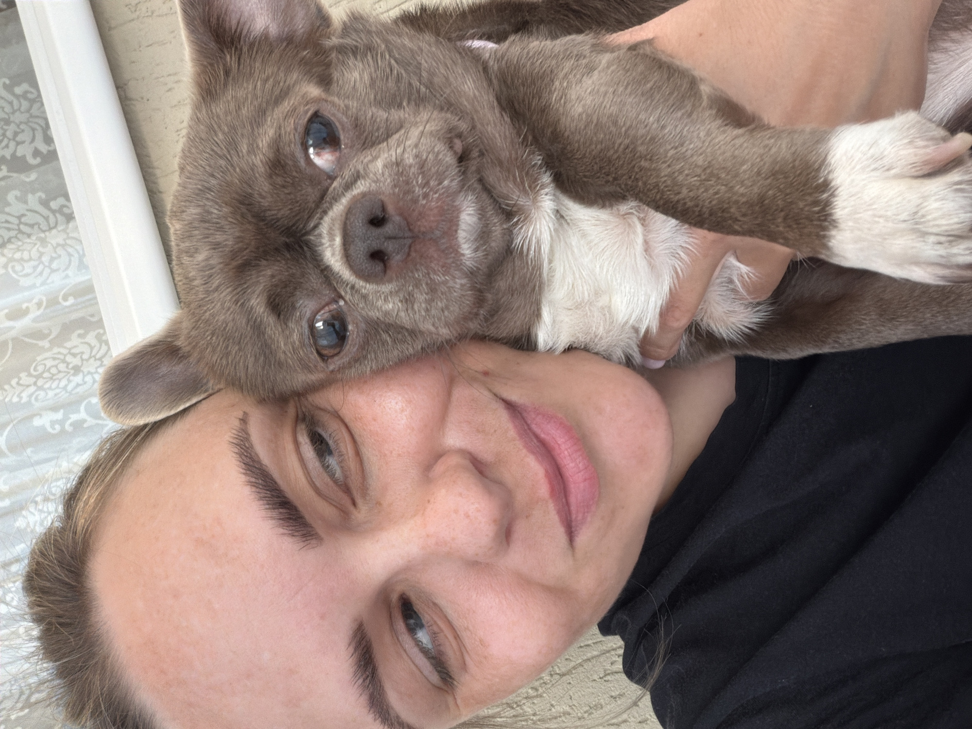 A selfie shows a woman with light skin and brown hair pressed against the head of a gray and white dog with a wrinkled face. The woman is wearing a black shirt and looking at the camera. The dog is being held by the woman. The background is a bed with a light colored patterned sheet. It is not possible to determine the location of the image to be Votkinsk, Russia.