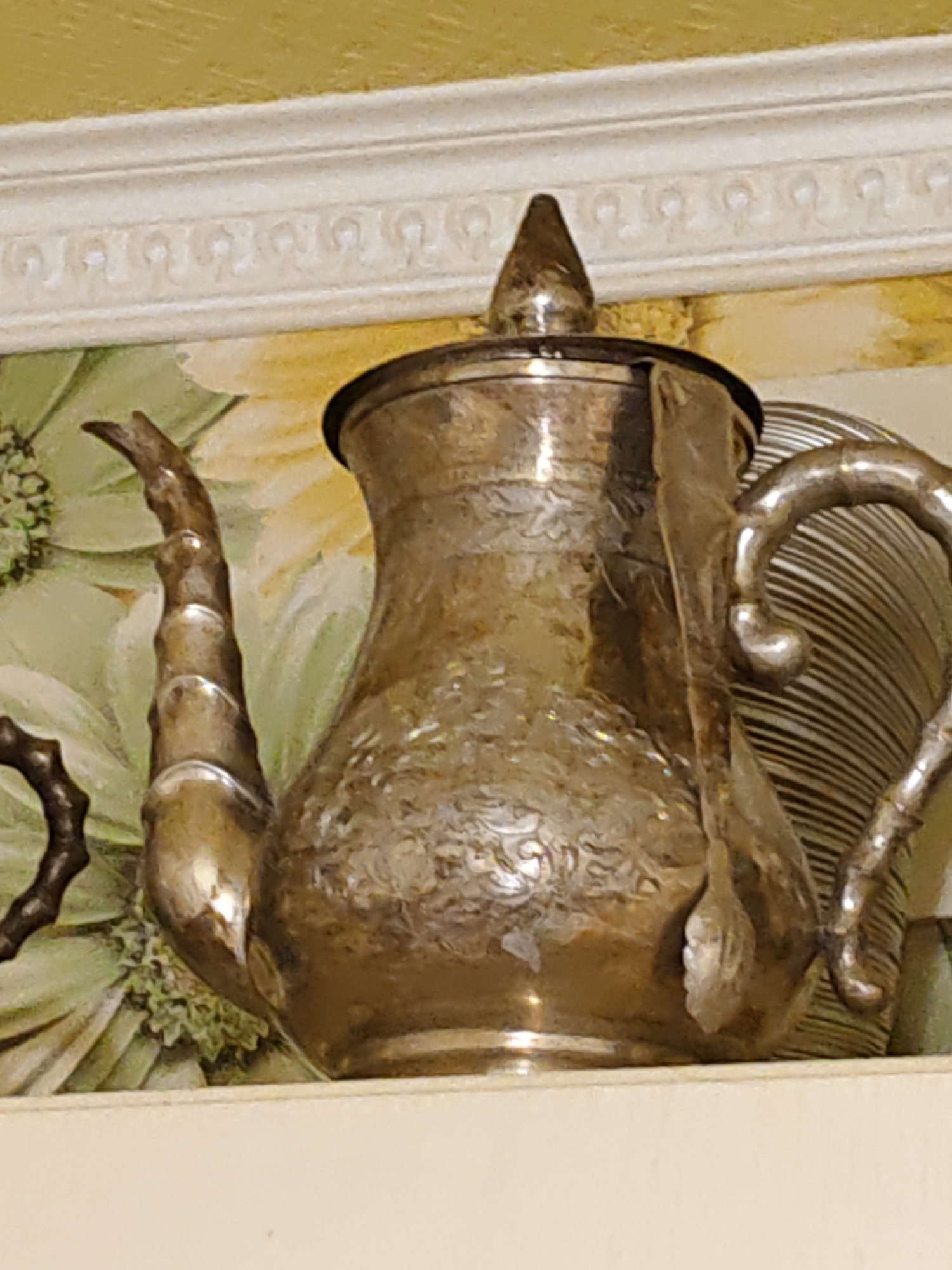 The image shows a close-up of an ornate, metallic teapot with intricate engravings. The teapot is silver or brass colored and has a curved spout and a detailed lid. It is positioned in front of a background that appears to be a floral print or painting. Above the teapot is a decorative white molding with a repeating pattern. The scene is lit in a way that highlights the metallic sheen of the teapot. There is no indication of the city of Rostov, Russia, or any specific environmental details that would suggest a location. The image is focused on the object itself, providing no context about its surroundings beyond the immediate background and decorative elements.