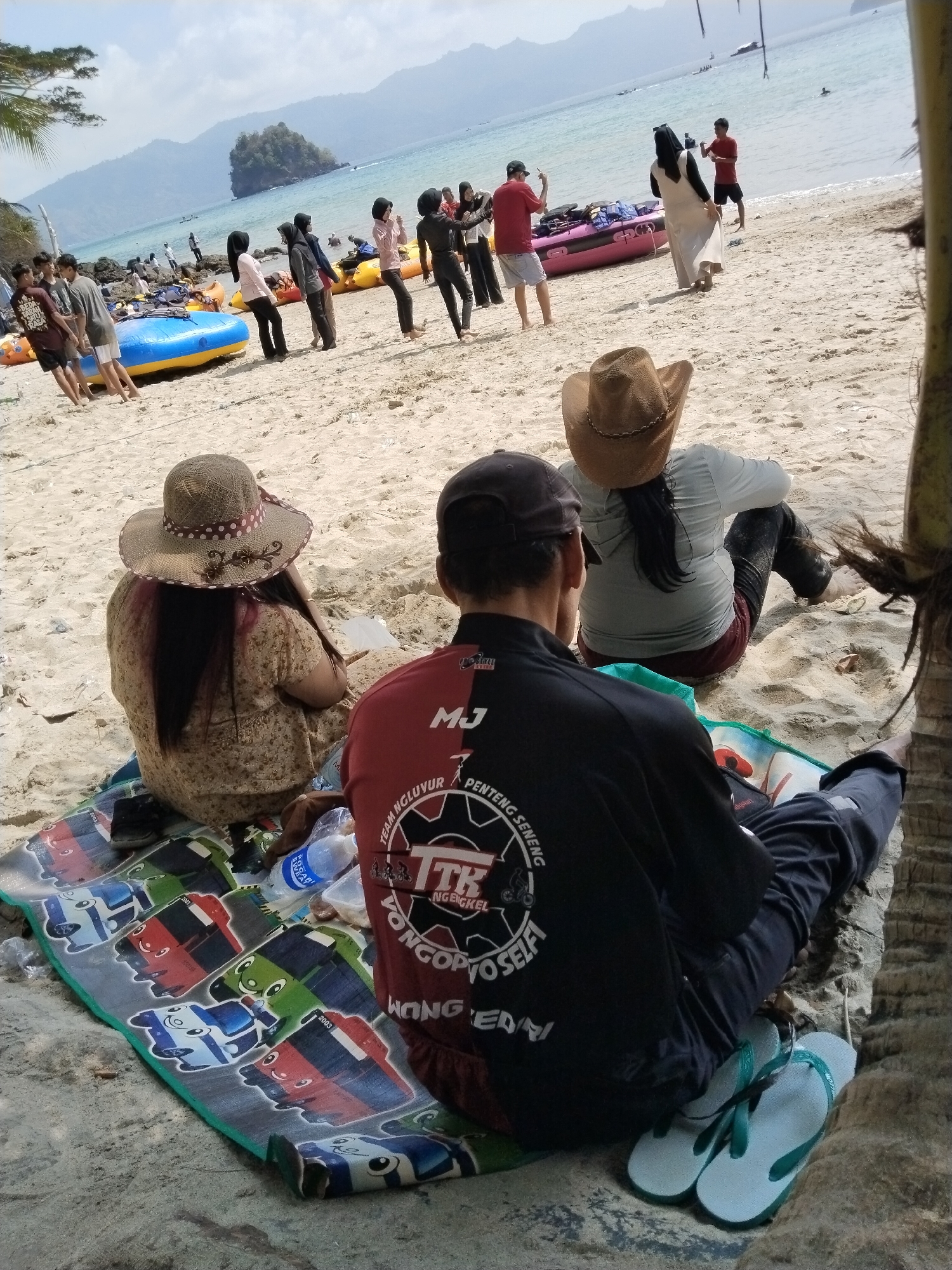 The image depicts a sandy beach scene in Prigi, Indonesia, with a view of the ocean and hills in the background. In the foreground, three individuals are seated on a colorful mat. Two of them are facing away from the camera, one wearing a black shirt with text and graphics and dark pants, and the other wearing a light-colored shirt and dark pants, with a cowboy hat. The third person, also facing away from the camera, is wearing a floral-patterned dress and a straw hat.
Further back on the beach, several other people are visible, some standing and others interacting with inflatable rafts. The water appears calm, and boats can be seen in the distance. The overall atmosphere suggests a recreational outing at the beach.