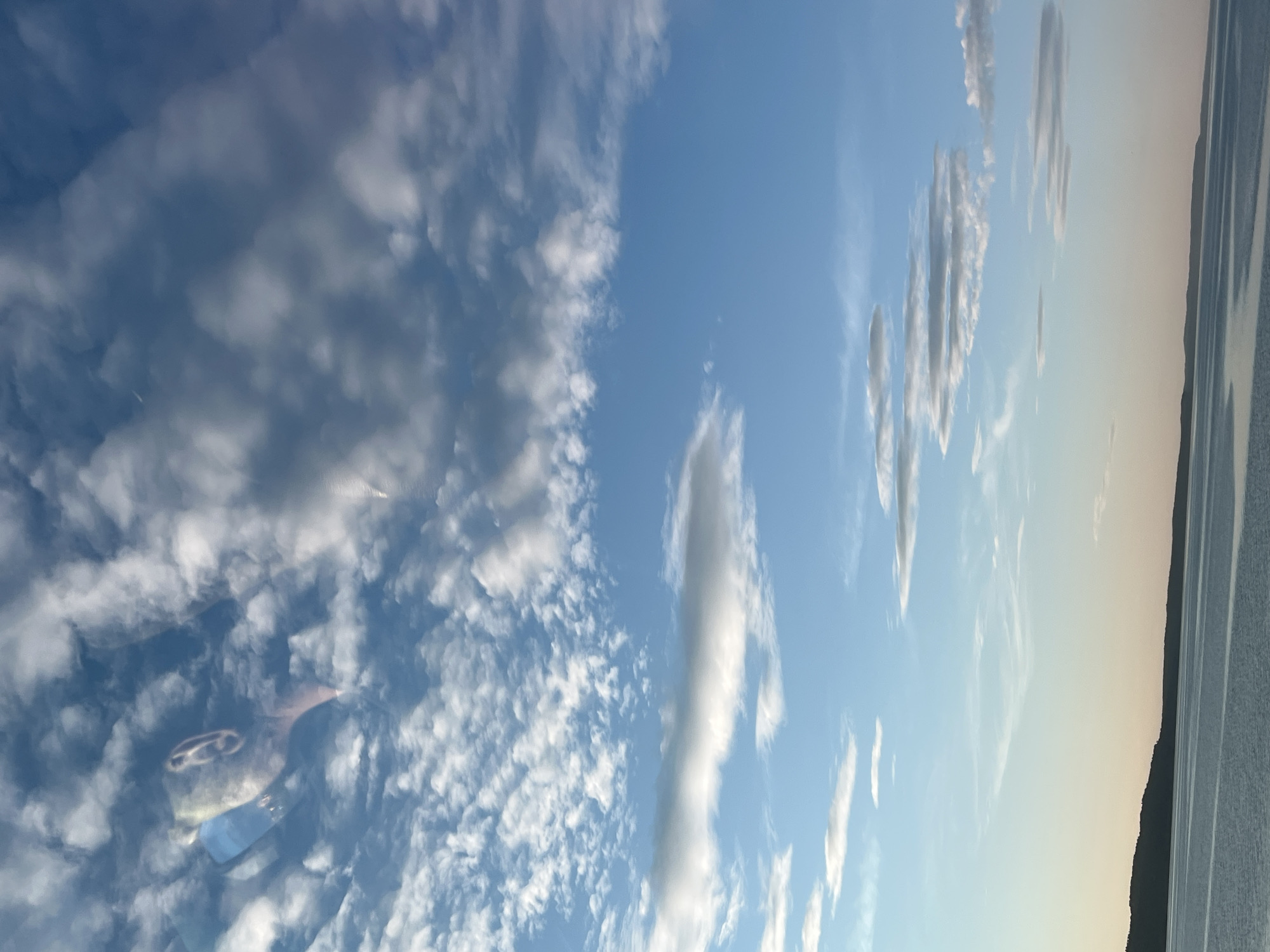 The image presents an outdoor landscape captured through a reflective or translucent surface, revealing a wide body of water in the foreground and a distant, tree-lined landmass under a partly cloudy sky. The lighting conditions suggest either early morning or late afternoon, with the sky transitioning from a soft blue overhead to a faint warm orange-pink at the horizon line.
The upper portion of the sky is populated by a mixture of white and grey clouds, ranging from denser, lumpy formations in the upper left to more scattered and elongated clouds across the middle and right sections. A faint, thin linear trace, possibly a contrail, is discernible in the upper mid-right.
Dominating the upper left quadrant is a distinct, semi-transparent overlay or reflection, indicating the photograph was taken through a window. This overlay reveals internal elements, including a blurred, rounded object resembling part of a vehicle's interior (such as a steering wheel or headrest), a faint, indistinct outline of a human face or head (possibly wearing eyewear), and a blue, amorphous shape.
The lower half of the image is dominated by a large body of water, likely the Votkinsk Reservoir on the Kama River near Chaikovsky, Russia. The water surface is calm, displaying subtle ripples and reflecting the ambient light and sky in tones of blue-grey. A dark, undulating silhouette of land, indicative of a forested shoreline, forms the distant horizon, separating the water from the lower expanse of the sky.