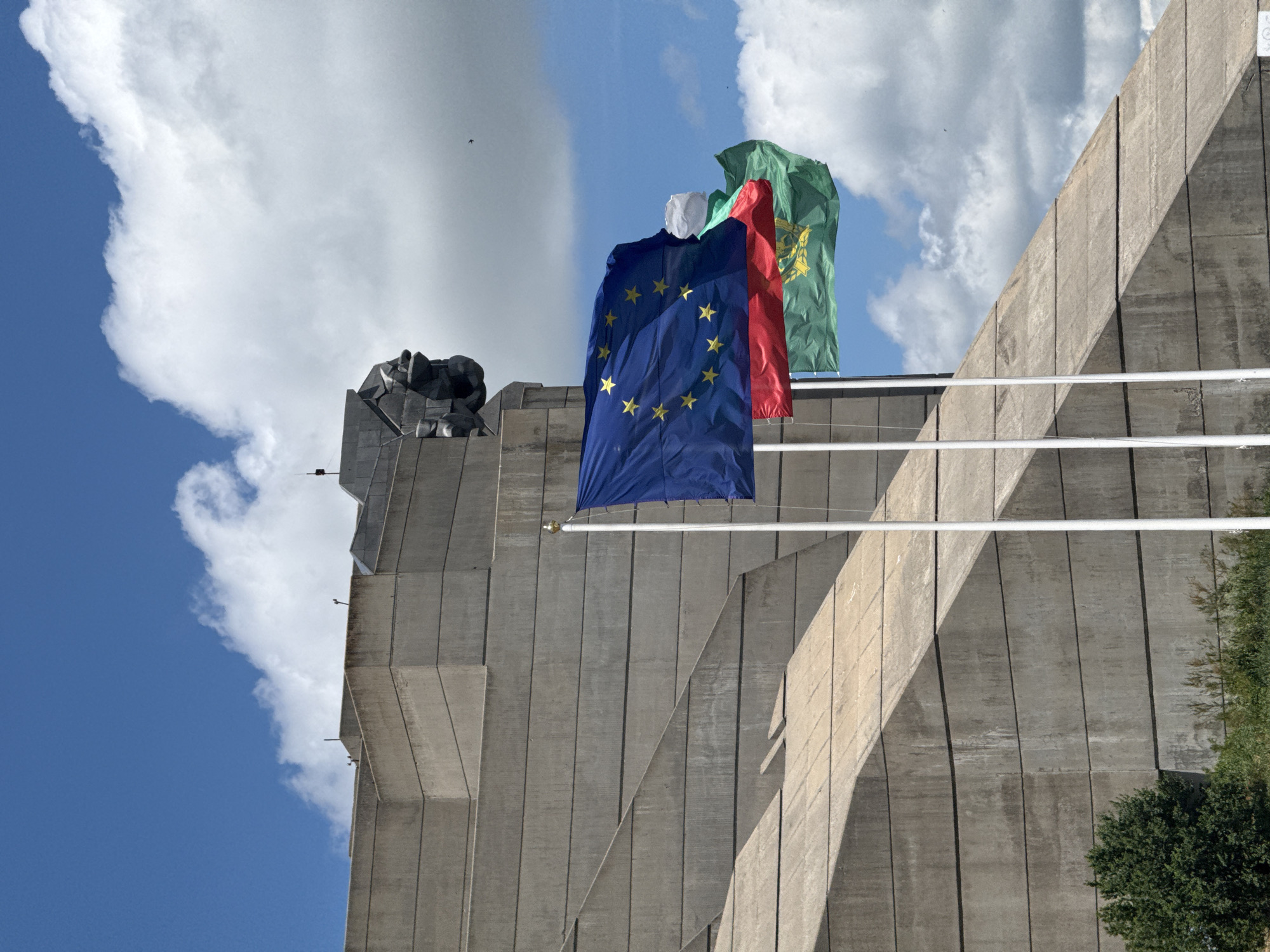 The image shows the Monument to the Declared of Bulgarian State in Shumen, Bulgaria, against a backdrop of a blue sky with white clouds. The monument is a large concrete structure with geometric patterns and sharp angles. In the foreground, three flags are flying on poles. The topmost flag is the flag of the European Union, with its blue field and yellow stars. Below it is the flag of Bulgaria, with its white, green, and red horizontal stripes. Partially obscured by the Bulgarian flag is another flag, predominantly green, which appears to be the flag of the Balkan region or a historical Bulgarian flag. The monument itself features a large, abstract sculpture of a lion's head on its upper portion, symbolizing Bulgaria. Lush green trees and vegetation are visible at the base of the monument. The lighting suggests it is daytime, with sunlight casting shadows on the concrete structure.