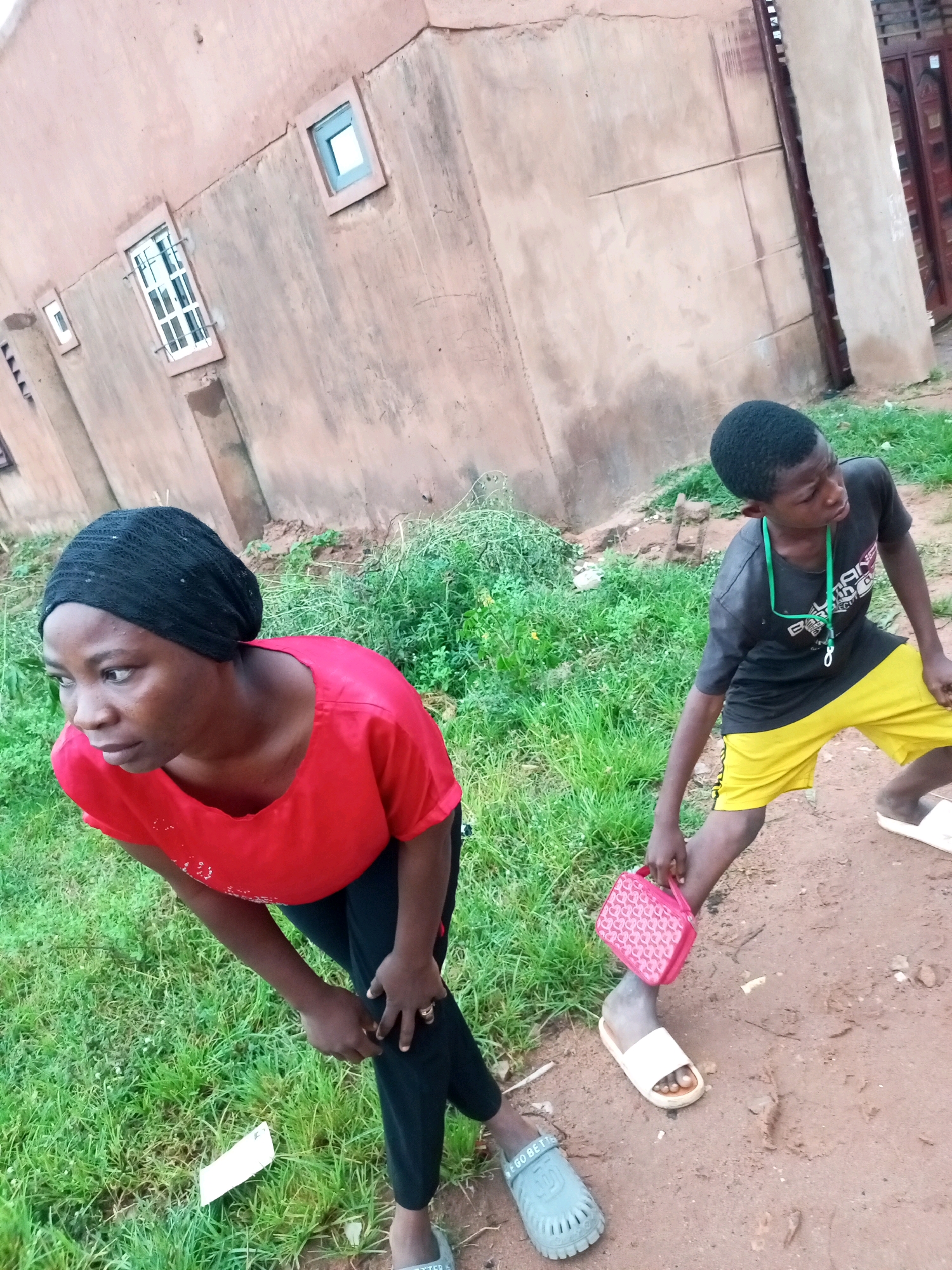 In Tempure, Nigeria, a young Black male wearing a black t-shirt with a green lanyard and yellow shorts is walking on a dirt path. He is holding a pink, patterned bag in his right hand. To his left, a young Black female wearing a red shirt and black headscarf is crouched down, looking to her left. She is wearing black pants and appears to be wearing rings on her left hand. The environment features a stucco building with windows and overgrown green grass.
