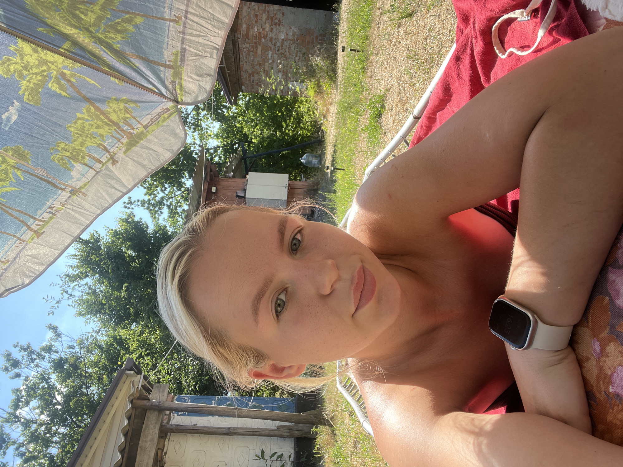 A young woman with blonde hair is lying on a red towel under a beach umbrella patterned with palm trees. She is wearing a black bikini top and a white smartwatch. The background shows a yard with a brick building, a wooden structure, and trees under a clear blue sky. The time displayed on the watch is 16:23.
