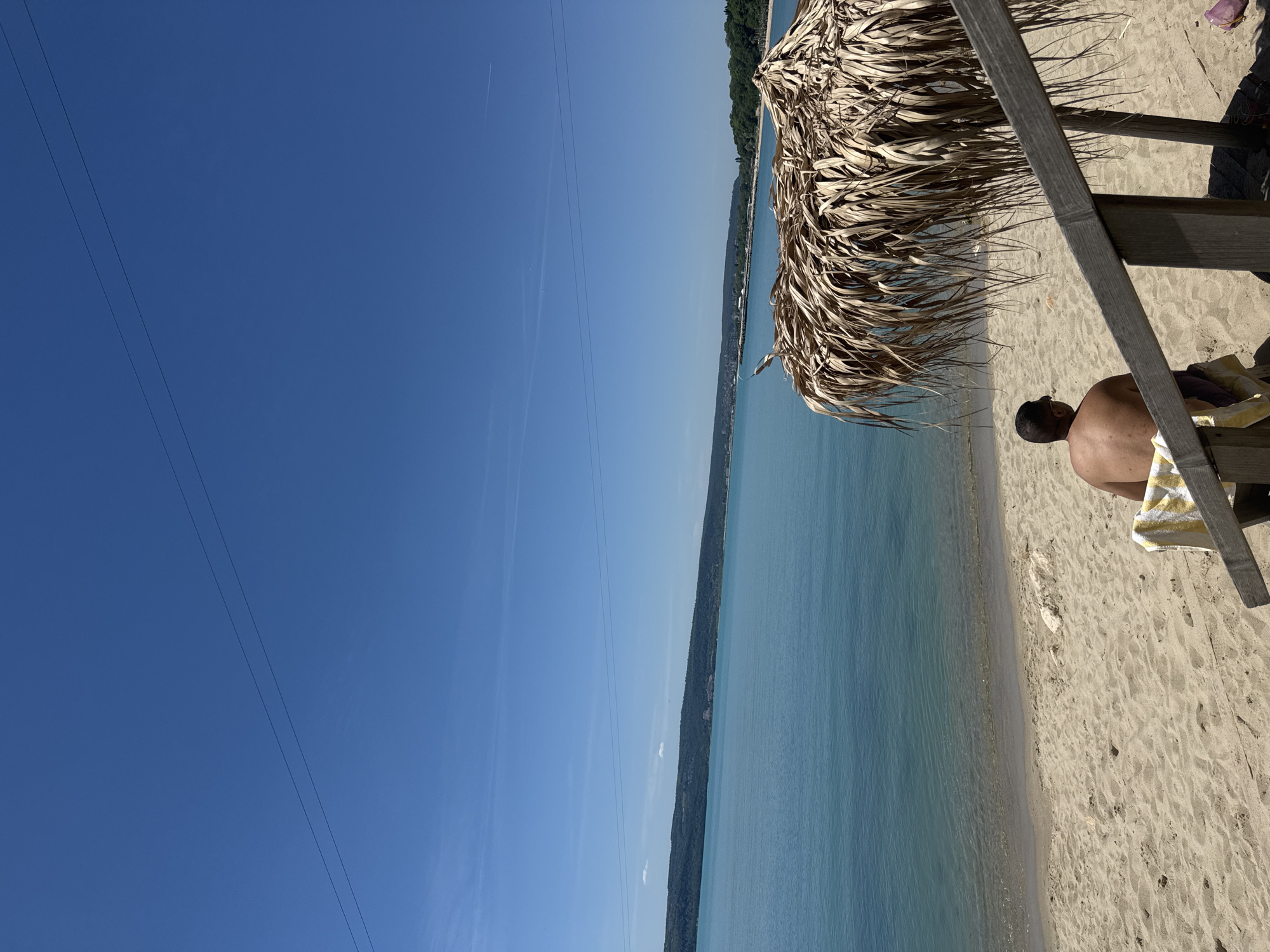 A photograph depicts a beach scene, likely in Varna, Bulgaria. The image is oriented with the sky on the left. The sky is clear, blue, and contains faint contrails. Power lines are visible across the upper left portion. A body of water, presumably the Black Sea, stretches along the middle. A coastline with dense trees is visible in the distance. A beach is present on the right. On the beach, a palapa-style umbrella or sunshade is constructed with dried palm fronds. A person is sitting in a beach chair on the sand, under the shade structure. The person has dark hair and a dark complexion. The chair is covered with a yellow and white striped towel. The beach is composed of light-colored sand.