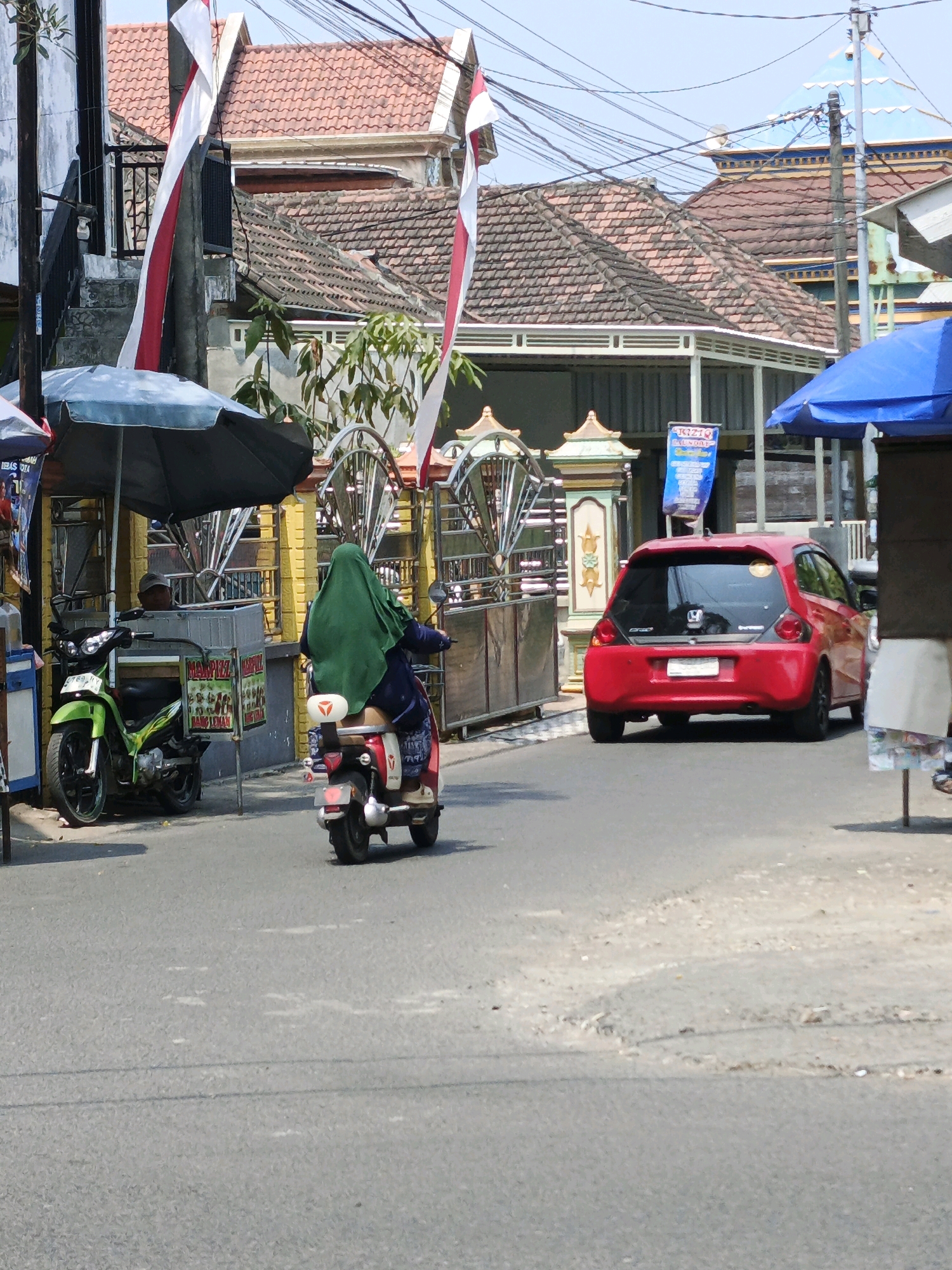 The image depicts a street scene in Ketegan, Indonesia, under a bright, clear sky. In the foreground, the asphalt road shows signs of wear with patches of lighter material. To the left, a green motorcycle is parked under a dark blue umbrella. A person wearing a cap is seated behind a counter or cart that displays food items with Indonesian text. A woman wearing a green headscarf and dark clothing rides a red scooter away from the viewer. She is traveling in the same direction as a red hatchback car visible in the middle ground of the image. The car has tinted windows and a license plate. Buildings with red tiled roofs and white trim line the street, with some structures featuring decorative elements. Several power lines crisscross the sky above. Red and white flags are displayed on poles near the buildings. The overall atmosphere suggests a typical day in a populated area of Indonesia.