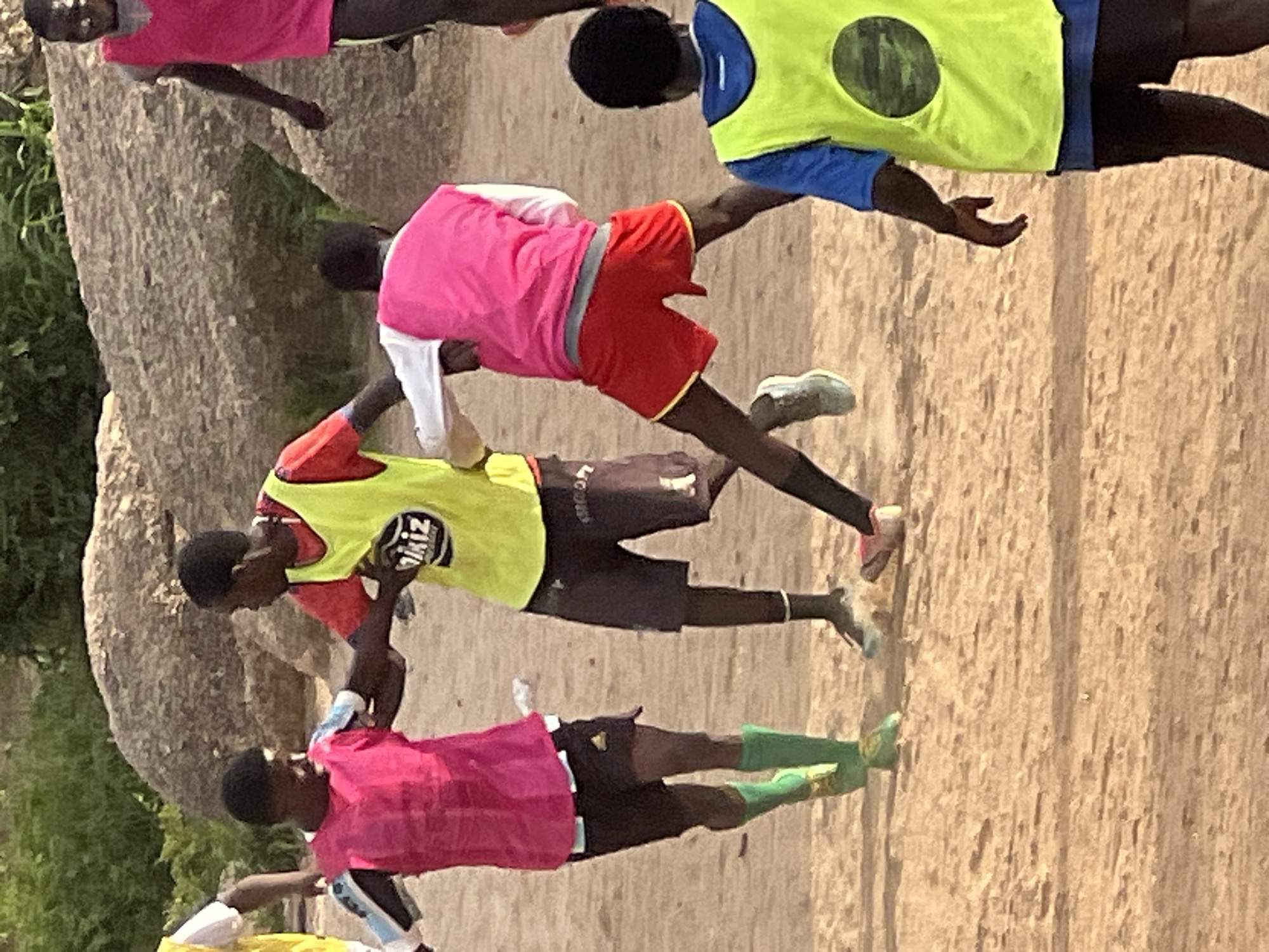 This image depicts a group of young people engaged in a game of football on a dirt field in Jos, Nigeria. The scene is characterized by the dry, sandy ground and a backdrop of large rocks and sparse vegetation. Several individuals are wearing brightly colored athletic vests, primarily pink and yellow, over their sportswear. One person in a pink vest and black shorts, wearing green socks, is in the foreground to the left. To his right, another individual in a red shirt and yellow vest, also in black shorts, appears to be actively participating in the game. Another person in a pink vest and red shorts is visible to the right, also seemingly involved in the play. In the background, a person in a pink vest is partially visible. The focus is on the dynamic activity of the game, with players in motion and interacting with each other on the dusty field. The environment suggests an outdoor recreational area within the city of Jos.