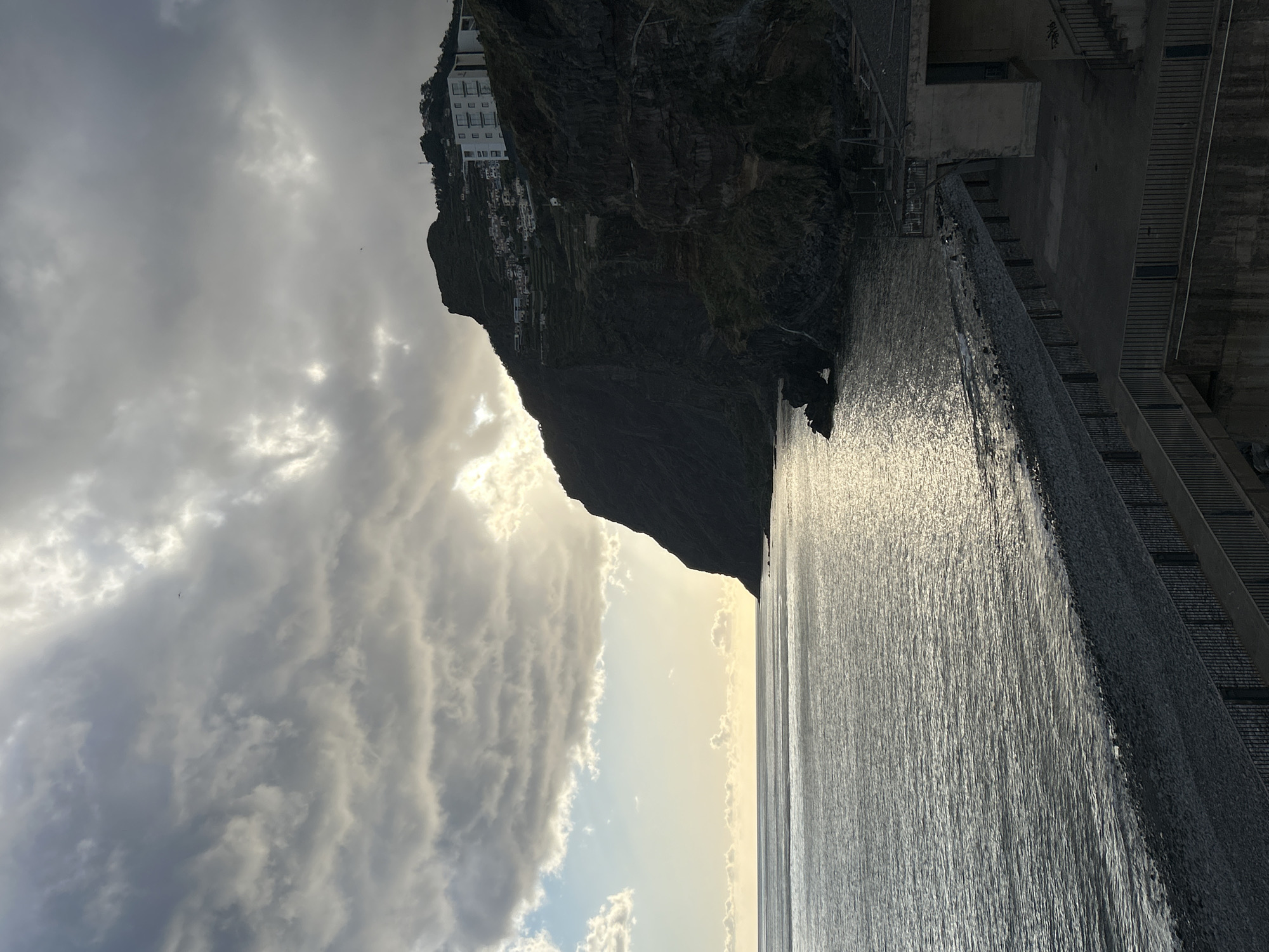 The image depicts a landscape in Câmara de Lobos, Portugal. The scene includes a steep, dark cliff on which buildings are perched, a body of water reflecting light, and a cloudy sky. A large structure, possibly a building or wall, is visible on the right side. The interaction between the sunlight and clouds creates a contrast with the darker cliff and water.