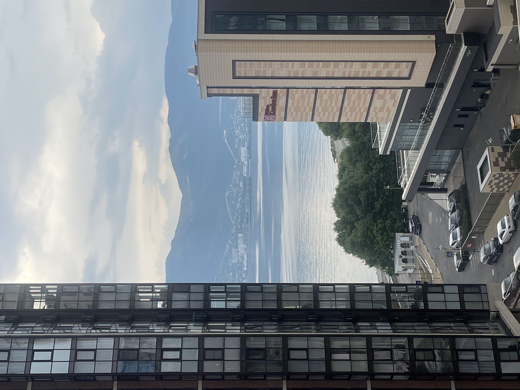 The image captures a panoramic view of the Novorossiysk embankment and its surroundings, likely taken from a high vantage point within one of the residential buildings. Dominating the left side of the frame is the modern facade of a tall apartment building, characterized by numerous windows reflecting the overcast sky. To the right, the Hilton Garden Inn hotel is visible, its beige and dark facade distinguished by balconies and tinted windows.
Beyond the buildings, a calm sea stretches out, meeting a distant coastline dotted with structures and a hilly landscape that fades into a hazy blue mountain range under a cloudy sky. The foreground on the right reveals a paved area with parked cars and a small coffee kiosk, indicating an active urban environment. The overall impression is one of a seaside city under a subdued, possibly early morning or late afternoon light.