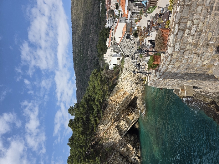 The scene is located in Medinski Krš, Montenegro, under a clear blue sky with scattered white clouds. The left mid-ground features a steep, stratified rock face with a prominent cave entrance at water level, topped by evergreen trees. Below, the water is a deep turquoise. The foreground is defined by a stone retaining wall, behind which an elevated outdoor dining terrace is visible. This terrace contains numerous dark-colored tables and chairs, two black decorative lampposts, and multiple terracotta planters holding green foliage. Beyond the terrace, a cluster of white and stone buildings with red-tiled roofs constitutes a coastal settlement. The background consists of densely vegetated hills. Two individuals are present on the terrace: one in dark attire is seated at a table, and another in a blue garment is partially visible further right. A blue object is visible at the water's edge on the far right.