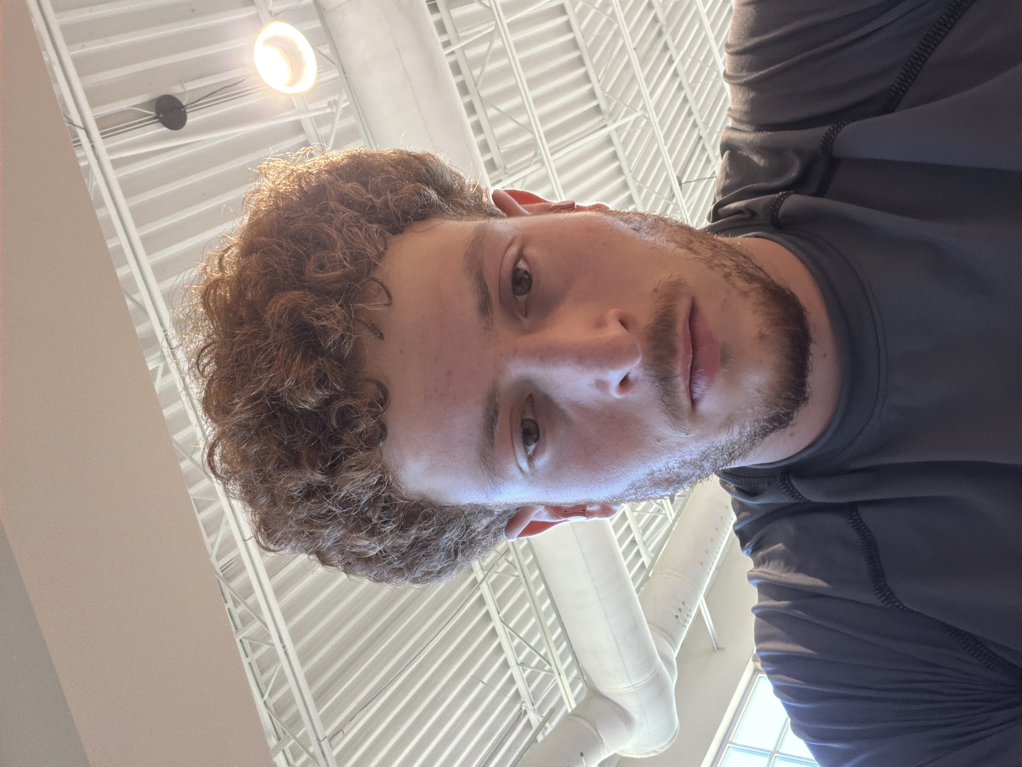 A low-angle, medium shot captures a young Caucasian male with curly, auburn hair and a light beard from the shoulders up. He is wearing a dark gray, athletic-style shirt with visible black stitching. The background consists of a white, corrugated ceiling with exposed beams and ducts, illuminated by fluorescent lighting and a bright, circular fixture. A large window with diffused sunlight is visible on the left side, hinting at an indoor, possibly commercial or recreational, setting. The man's face is turned slightly downwards, with his eyes looking towards the camera, and his expression appears neutral. The image appears to be taken in an indoor space, potentially a gym or a large open-plan office, within the city of Beachwood, United States of America.