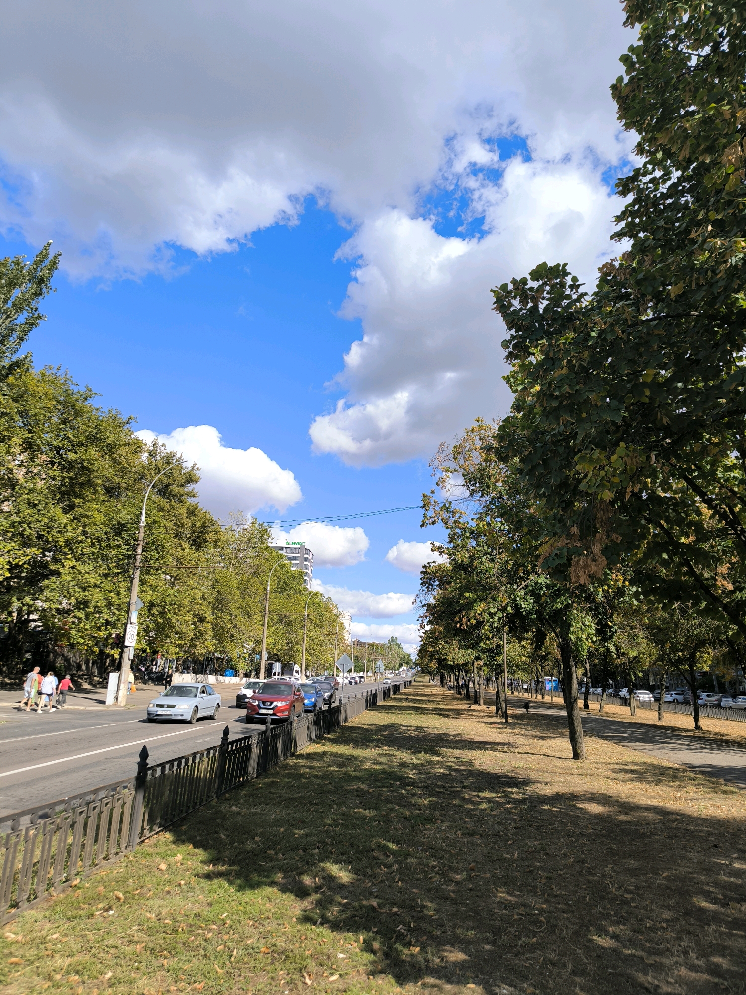 The image depicts a sunny day on a wide, tree-lined avenue in Mykolaiv, Ukraine. The sky is a vibrant blue, dotted with fluffy white clouds. On the left side of the avenue, a road with multiple lanes carries traffic, including several cars and a few pedestrians walking on the sidewalk. The trees on the left are lush and green, casting shadows on the road. On the right side of the avenue, a long row of trees with green and yellowing leaves lines a grassy median, with a black metal fence separating it from a sidewalk and more parked cars in the distance. In the background, a modern multi-story building with the "Invest" logo is visible. The overall scene suggests a typical urban environment on a pleasant day.
