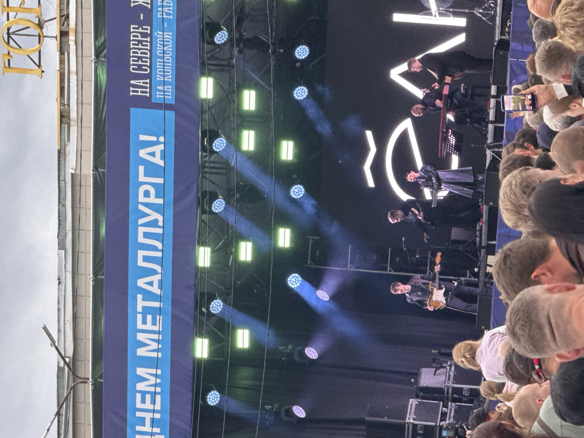 A large outdoor concert stage is set up, with a band performing in front of a dark curtain backdrop and a brightly lit logo. In the foreground, a crowd of people is gathered, watching the performance. The stage features several musicians, including a guitarist in a black leather jacket and jeans playing an acoustic guitar, a keyboardist in a dark trench coat, and another musician playing a keyboard. The backdrop has large white lettering, part of which is visible. Overhead, a rig of stage lights is suspended, with blue and green lights casting beams onto the stage and into the audience. A banner across the top of the stage reads in white letters "НЕМ МЕТАЛЛУРГА!" and "НА СЕВЕРЕ - Ж", and "НА КОЛЬСКОЙ - РАБ". In the upper right corner of the image, a large golden letter "Г" is visible, suggesting it might be part of a larger sign. The overall scene depicts a lively outdoor music event.