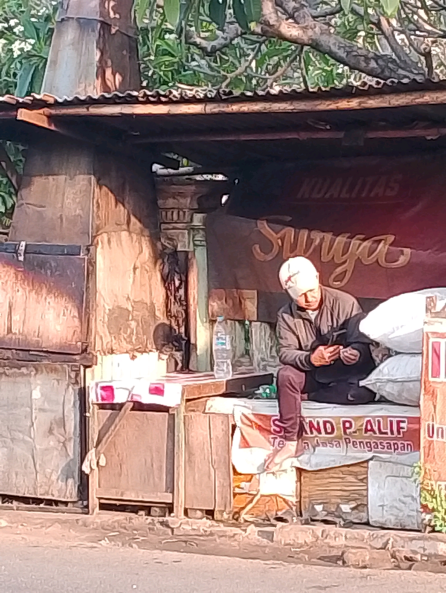 A man wearing a white cap and a grey jacket sits on a wooden bench under a corrugated metal roofed shelter. He is looking down and appears to be holding something in his hands. In front of him are several white sacks. Behind him, a banner with red lettering and the word "SURGA" in cursive script is visible. To the left of the man, a plastic bottle of water sits on a wooden surface. The shelter appears to be a small roadside stall in Probolinggo, Indonesia, as indicated by the signage in Indonesian, "SAND P. ALIF" and "Jasa Pengasapan." The ground in front of the stall is paved and a tree with green foliage is visible in the background.