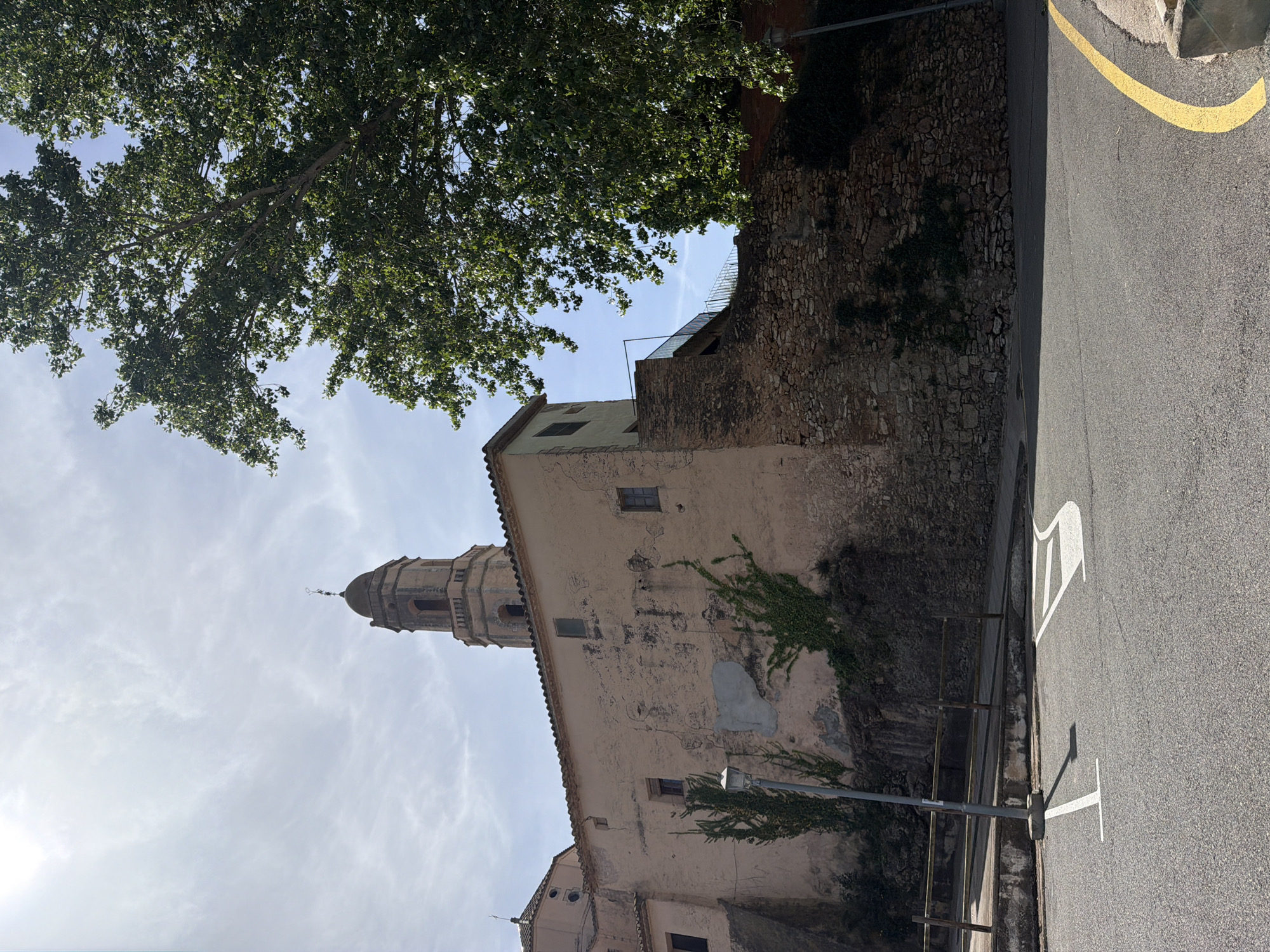 The image captures a view of the church in Bonastre, Spain. The church, featuring a prominent bell tower with a dome-shaped top, stands next to a stone wall partially covered with vegetation. A section of street runs alongside the wall, with a yellow line and painted white markings on the asphalt. Overhanging from the left is a large tree with dense foliage. The sky is visible behind the tree and the church, marked with wispy clouds. The lighting suggests it is daytime. A street lamp is positioned between the wall and the street.