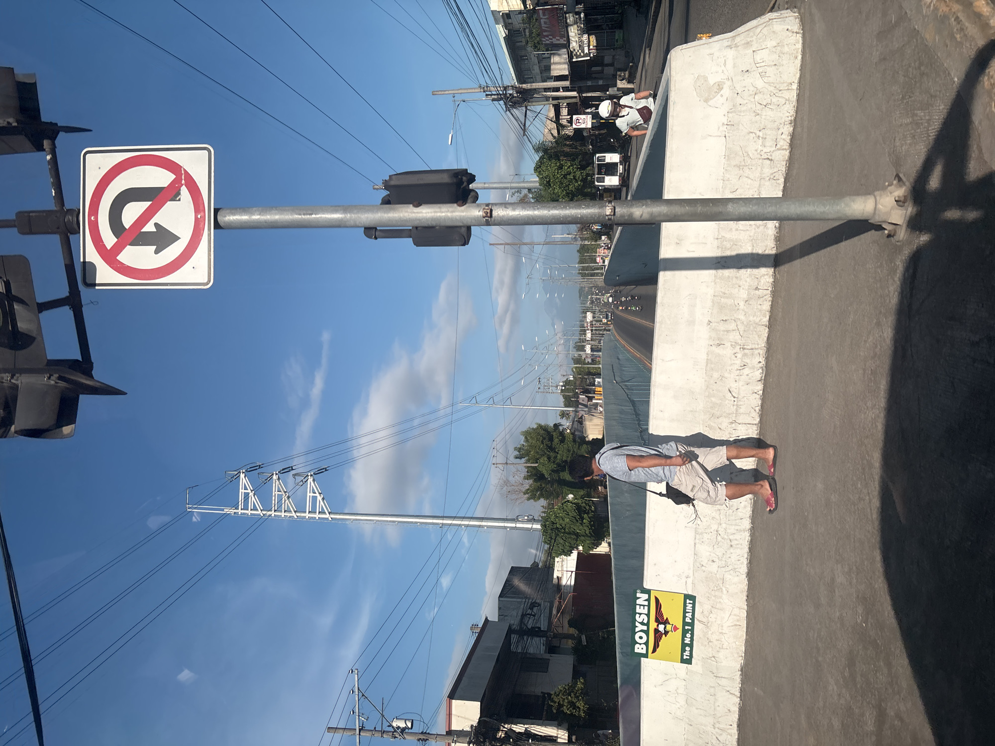 The image depicts a street scene in Davao City, Philippines, under a clear blue sky with scattered clouds. In the foreground, a man walks along a concrete pedestrian overpass, wearing a gray t-shirt, beige shorts, and flip-flops. He carries a dark bag over his shoulder. Adjacent to him on the overpass wall is a yellow sign with a black logo and text that reads "BOYSEN" and "The No. 1 PAINT".
A metal pole supporting traffic signals and a "no U-turn" sign stands prominently on the overpass. The sign features a red circle with a diagonal line through a black U-turn arrow pointing left, indicating that U-turns are prohibited.
Beyond the overpass, a road stretches into the distance, with vehicles including cars, motorcycles, and a white van visible. Power lines crisscross the sky, supported by numerous utility poles. Buildings line the sides of the road, some appearing to be commercial establishments. The overall environment suggests an urban setting with ongoing traffic and infrastructure.