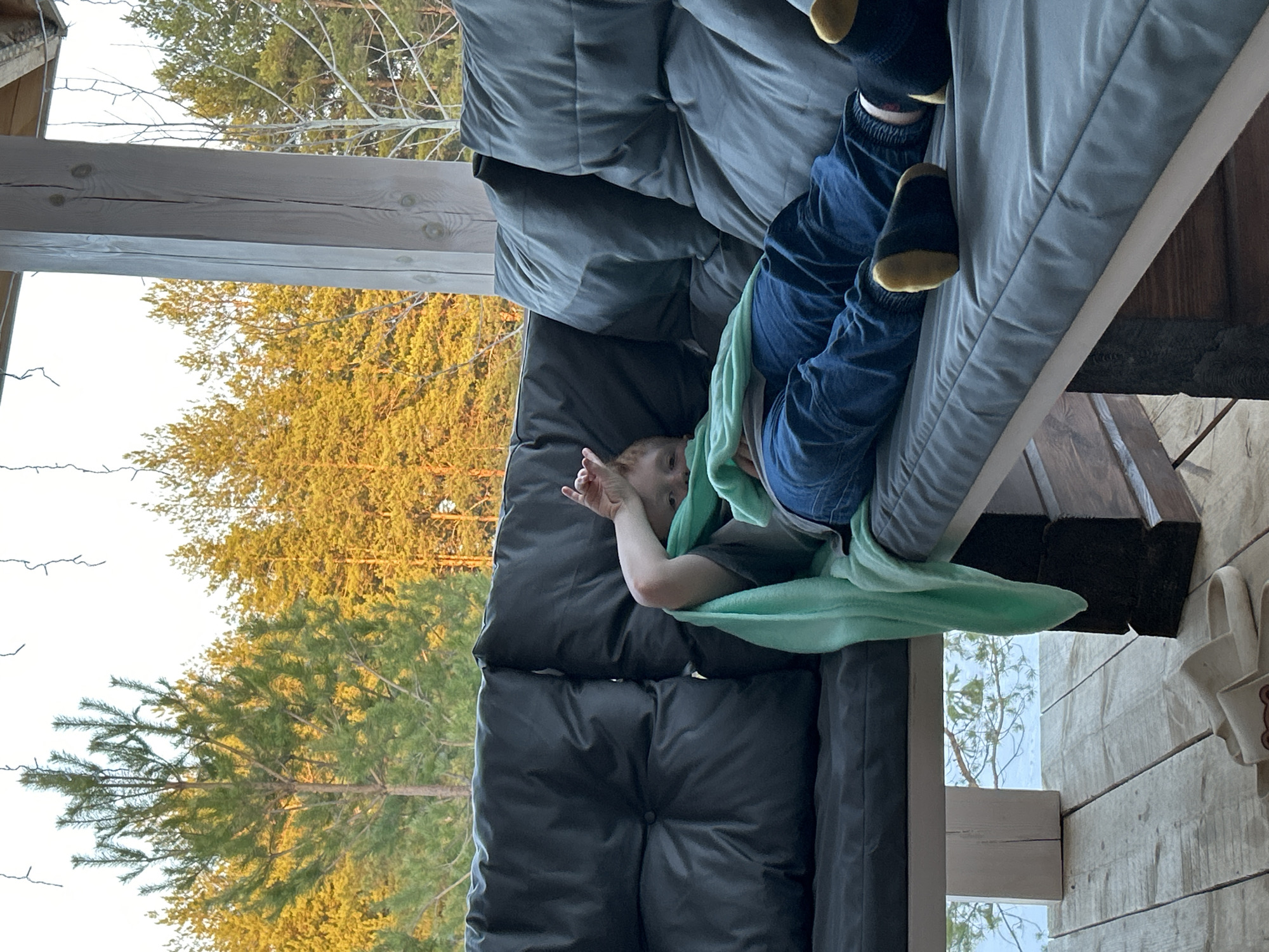 A child is reclining on a gray cushion-like structure indoors, covered partially by a light green blanket. They wear blue jeans and black socks with yellow accents at the toes and heels. Outside the window, tall green trees are visible. The photo was taken in Chaikovsky, Russia.