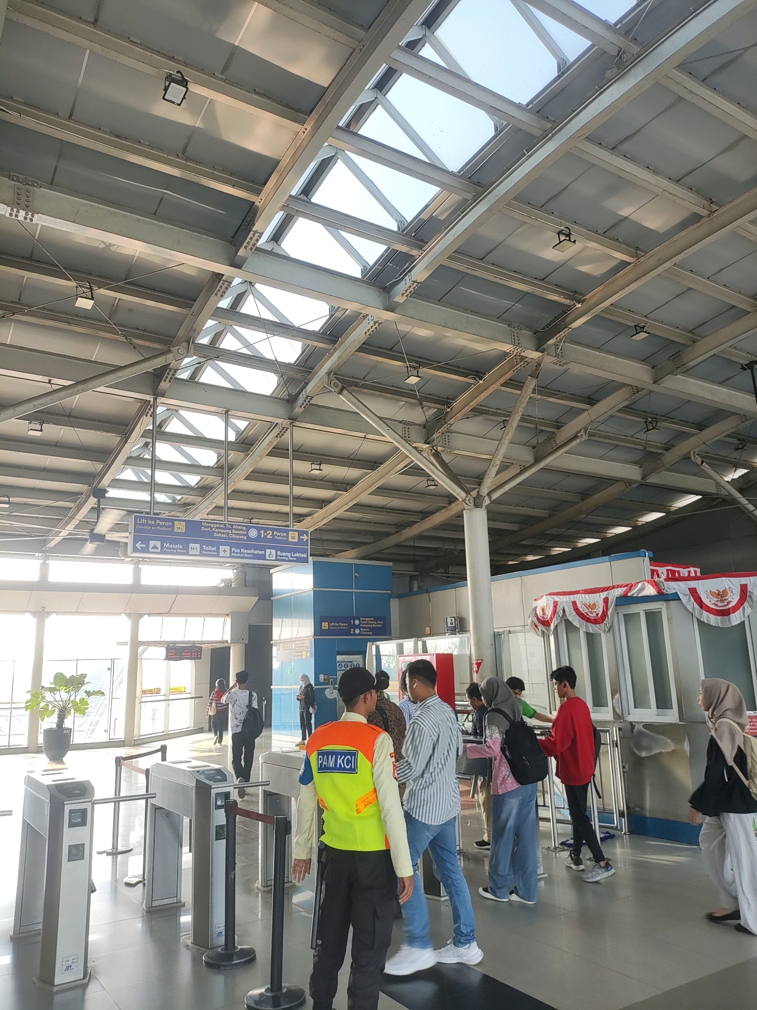 The image depicts an indoor area, likely a train station, with a high, exposed metal ceiling structure featuring skylights. In the foreground, a security guard wearing a bright yellow vest with "PAM KCI" text is standing to the right. In front of him are several automated ticket gates. A diverse group of people, including men, women, and young adults, are visible walking through or waiting near the gates. Some individuals are carrying bags. Signage is present, indicating directions and platform information. The environment appears clean and modern, with a tiled floor. The overall scene suggests a busy transportation hub. The specific location is identified as East Jakarta, Indonesia.