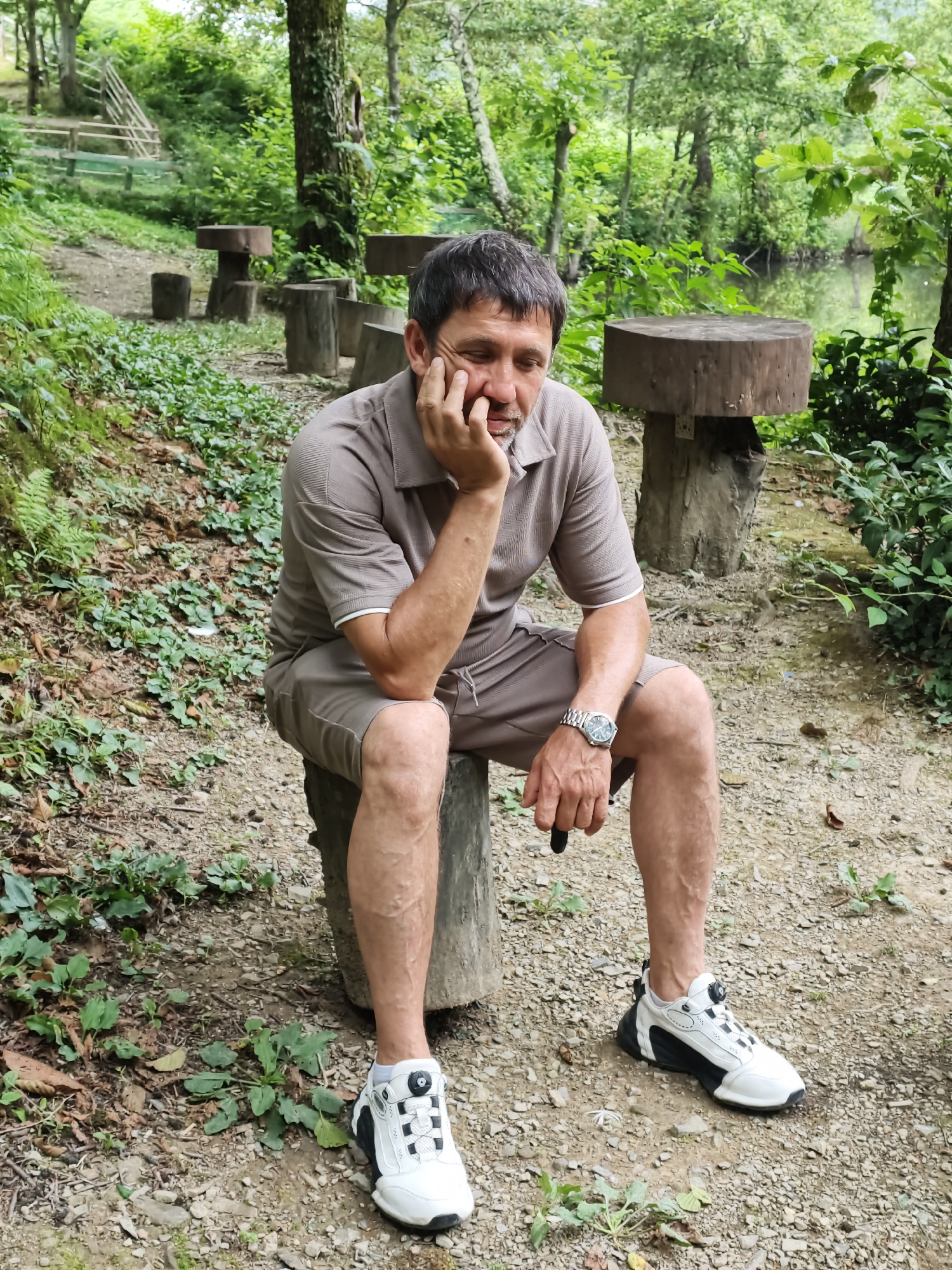An adult male with short, dark hair and stubble is sitting on a log in a wooded area. He is wearing a brown polo shirt and matching shorts, a silver wristwatch, and white and black athletic shoes. His right hand is resting on his chin, and his eyes are closed. In the background, there are more logs that appear to be used as seating, a wooden staircase, dense green foliage, and a body of water. The ground is covered with gravel and fallen leaves. The scene is set in Kalinovoye ozero, Russia.