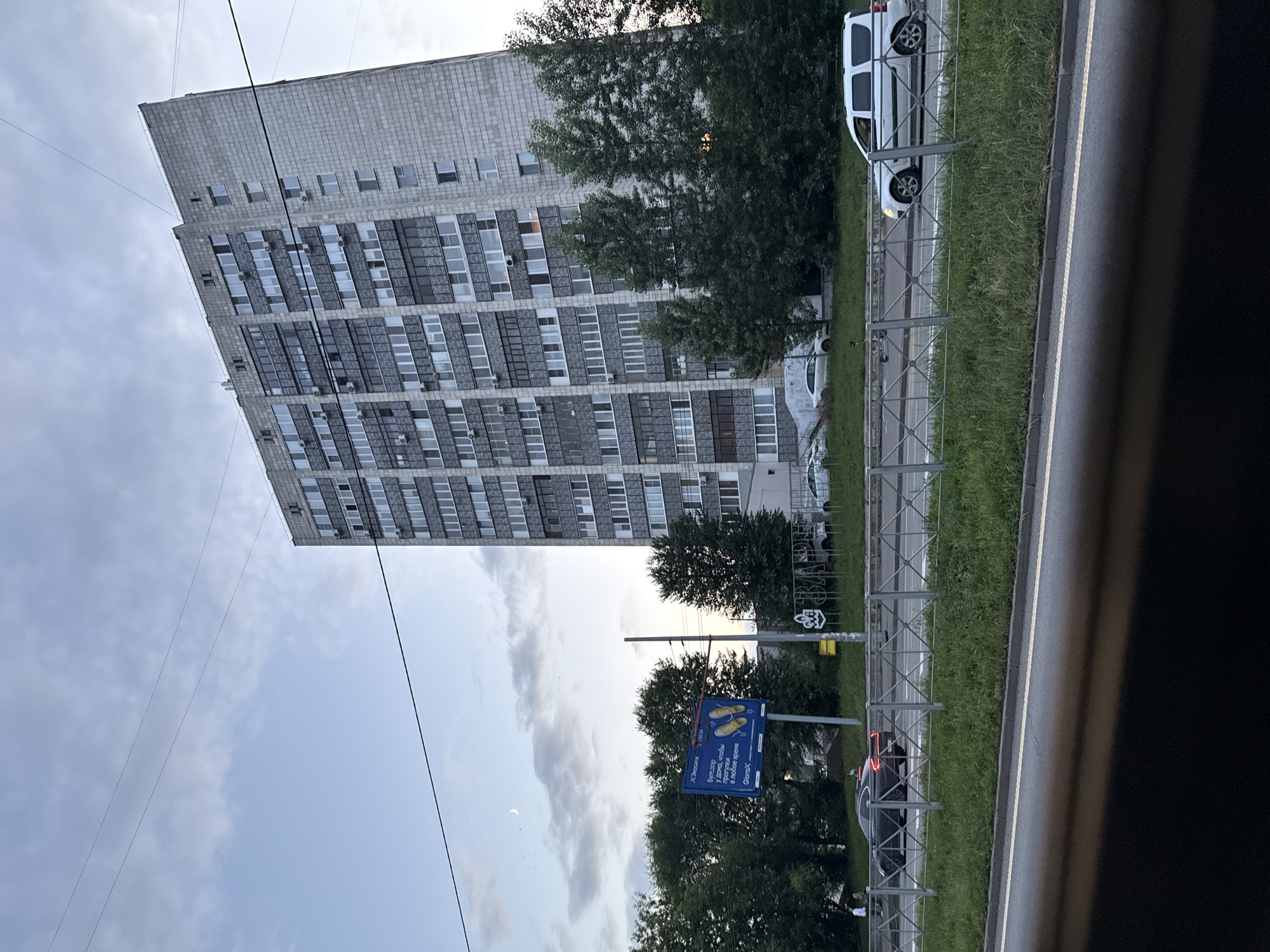 The image depicts a residential building in Kazan, Russia, with a gray facade and numerous windows. The building appears to be quite tall, possibly an apartment complex. In front of the building, there is a road with several cars, including a black sedan and a white SUV. A metal fence separates the road from a grassy area. There are trees behind the fence and in front of the building. The sky is overcast with some clouds, and a sliver of the moon is visible. A billboard with some text and images is also present on the left side of the image. The overall scene suggests an urban environment, likely a residential neighborhood.