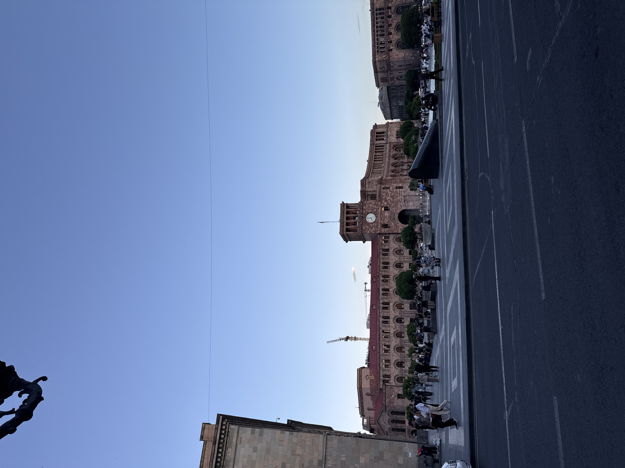 The image captures Republic Square in Yerevan, Armenia, from a street-level perspective. The square is flanked by several grand buildings, including one with a prominent clock tower. The architectural style is characterized by detailed facades and arched windows. A crane is visible, suggesting ongoing construction or renovation work. Pedestrians are present on the square, walking along the edges, and sitting on benches or ledges, and cars are parked, some next to benches with people. The road is marked with painted white lines and pedestrian crossings. The sky is clear and blue. A dark statue and building corners are visible in the foreground.