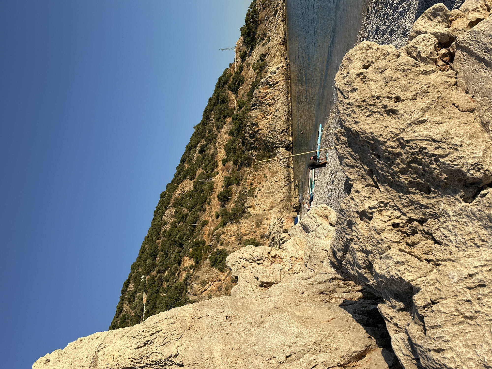 The image displays a serene coastal landscape under a clear blue sky, likely in Fethiye, Turkey. A steep, verdant hillside covered with dense green vegetation, including trees and shrubs, dominates the left side of the frame. Rocky outcrops are visible on the slope, and thin utility lines or cables traverse the hillside, with a tall metal structure, possibly an electricity pylon or communication tower, situated higher up.
At the base of the hillside, a calm body of water, presumably the sea, stretches horizontally across the mid-right portion of the image, meeting a narrow, light-colored pebble beach. On this beach, in the midground, a person wearing dark clothing and a hat is seen kneeling or sitting next to a light blue and white inflatable paddleboard or kayak. They are holding a long, slender wooden pole, suggesting preparation for or engagement in a water activity. Another person is partially visible further along the beach.
The foreground and right side of the image are characterized by large, textured, light-colored rocks. These rocks exhibit detailed weathering patterns and crevices, illuminated by warm, golden sunlight that casts distinct shadows, indicating either early morning or late afternoon. The overall scene conveys a peaceful atmosphere, highlighting outdoor recreation by the sea. No visible text is present in the image.