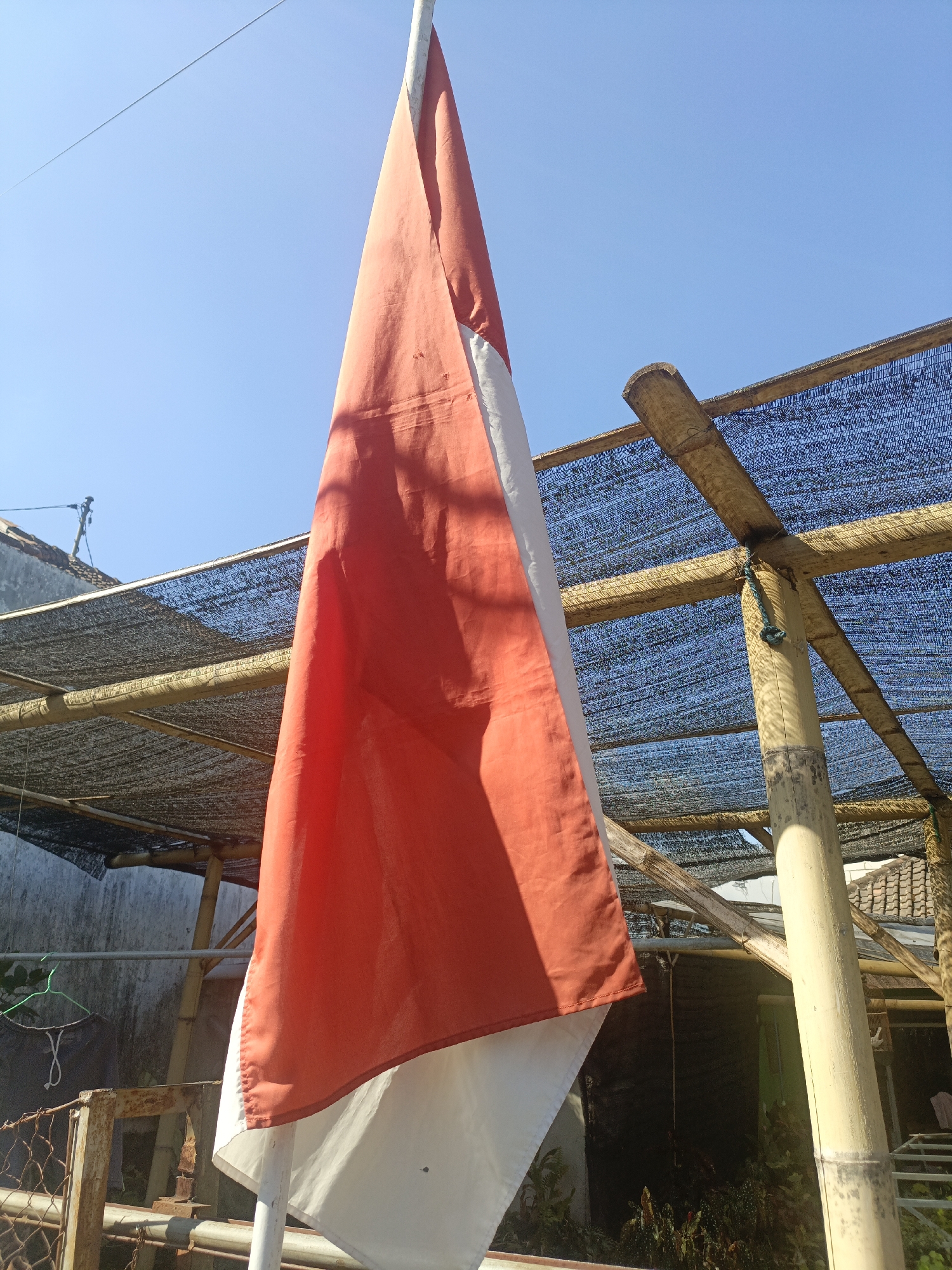 The image shows an Indonesian flag, red and white, hanging vertically against a clear blue sky. The flag appears to be made of fabric and is partially illuminated by sunlight, casting shadows on its surface. In the background, there is a structure made of bamboo poles and covered with a dark mesh material, suggesting it might be a shade or canopy. A light-colored wall and some foliage are visible beneath the structure. To the left, a metal fence and some utility poles are present. The scene is set outdoors under bright daylight.