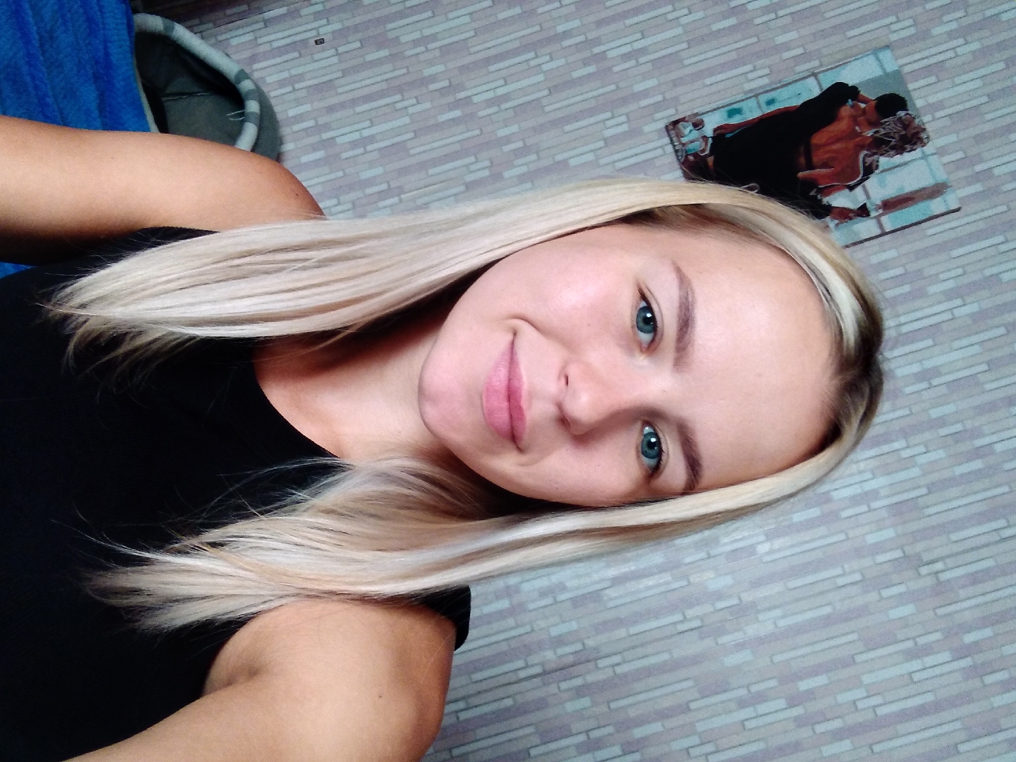 A young woman with blonde hair and blue eyes takes a selfie in what appears to be a bedroom. She is wearing a black top. Behind her, a picture of a couple embracing hangs on a wall with a patterned wallpaper. A blue blanket and a grey pet bed are visible in the background. The location is Radisne, Ukraine.