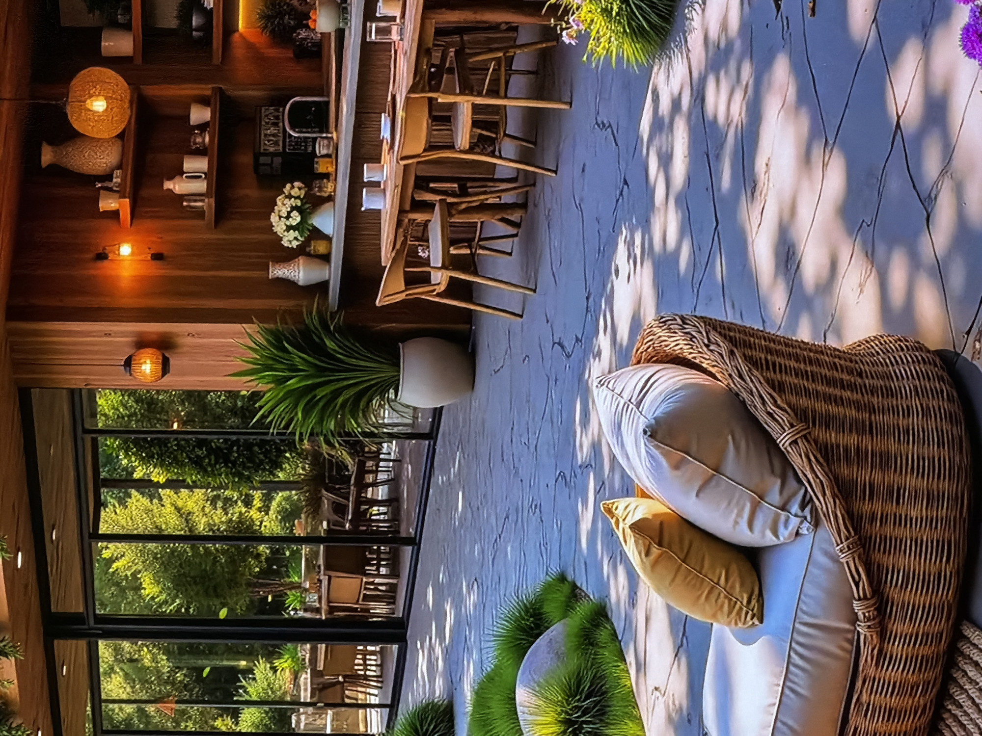 This image captures a bright and inviting indoor-outdoor setting, likely a restaurant, cafe, or a stylish private space, situated in Sofiivska Borshchahivka, Ukraine. The scene is illuminated by strong daylight, casting distinct dappled shadows from tree leaves onto the paved outdoor area, indicating a sunny day.
To the right, an outdoor lounge area features a comfortable wicker armchair furnished with light-colored cushions and two throw pillows, one light grey and one mustard yellow. The paving consists of light-toned, irregularly patterned slabs, possibly stone or concrete. Green ornamental plants, some with small purple flowers, are arranged around this seating area.
To the left, an interior space, likely a bar or a section of a dining room, is characterized by warm wooden walls and shelving. The shelves display various decorative items, including ceramic vases, pottery, and several small, glowing light fixtures, creating a cozy atmosphere. A long wooden table or bar counter is visible, accompanied by multiple wooden high-back chairs or stools, arranged for dining or casual gathering. A tall, leafy green potted plant stands near a large glass partition.
This glass partition offers a clear view of a lush green outdoor landscape beyond, filled with numerous trees and foliage. Through the glass, additional outdoor seating and tables can be seen, suggesting an extended dining or relaxation area that blends with the natural environment.
No people are visible in the image, leaving the space serene and unoccupied. The overall impression is one of tranquility, comfort, and a harmonious connection between the interior and the surrounding natural beauty. No discernible text is visible within the frame.