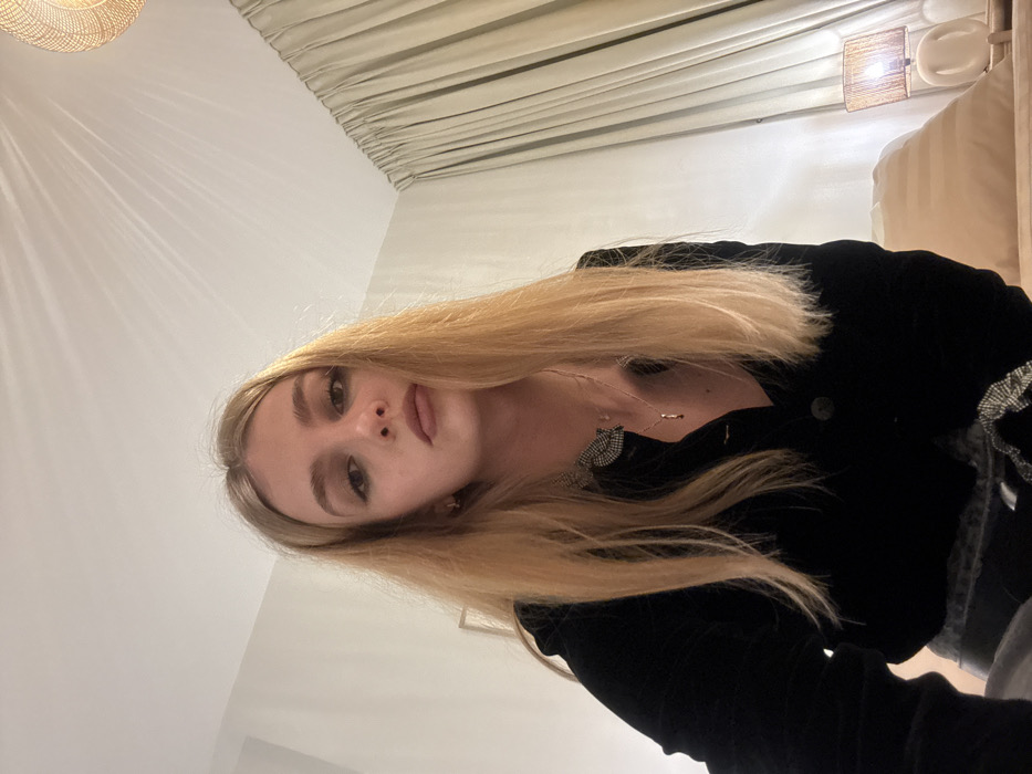 A fair-skinned woman with blonde hair poses for a selfie, her head tilted towards the right. She wears a black jacket, with a glimpse of a patterned top underneath. The background includes a white wall, draped curtains, and a wicker-style ceiling light. A bedside lamp and part of a beige couch are visible in the upper right corner.