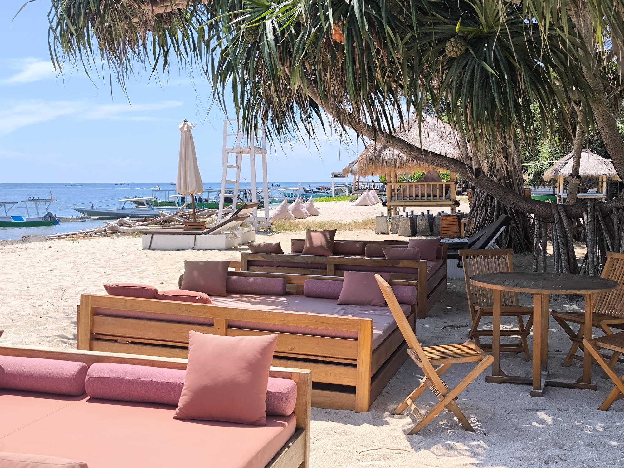 The image shows a beach scene in an unknown city in Indonesia. The foreground contains outdoor furniture, including lounge beds with pink cushions and a round table with wooden chairs. The beach is sandy, and the background includes a lifeguard stand, boats, and small huts with thatched roofs. The sky is blue. The scene is mostly deserted. Some of the image is partially obscured by the foliage of a tropical tree.