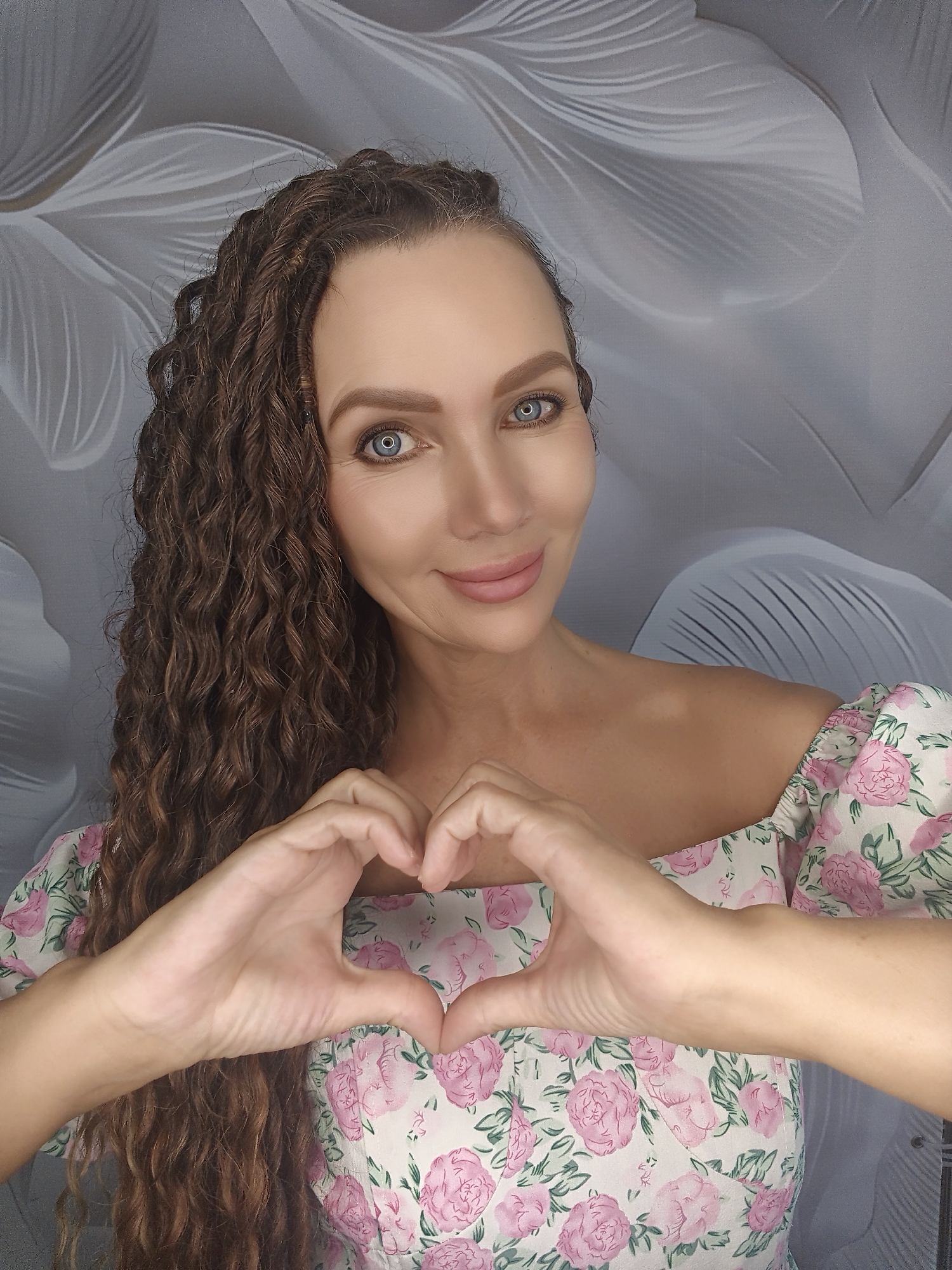 A medium shot captures a woman with long, dark curly hair, her eyes are blue and she has a gentle smile. She is wearing a floral-patterned off-the-shoulder dress. Her hands are clasped together in front of her chest, forming a heart shape. The background is a gray wall with a subtle pattern of white, feathery designs.