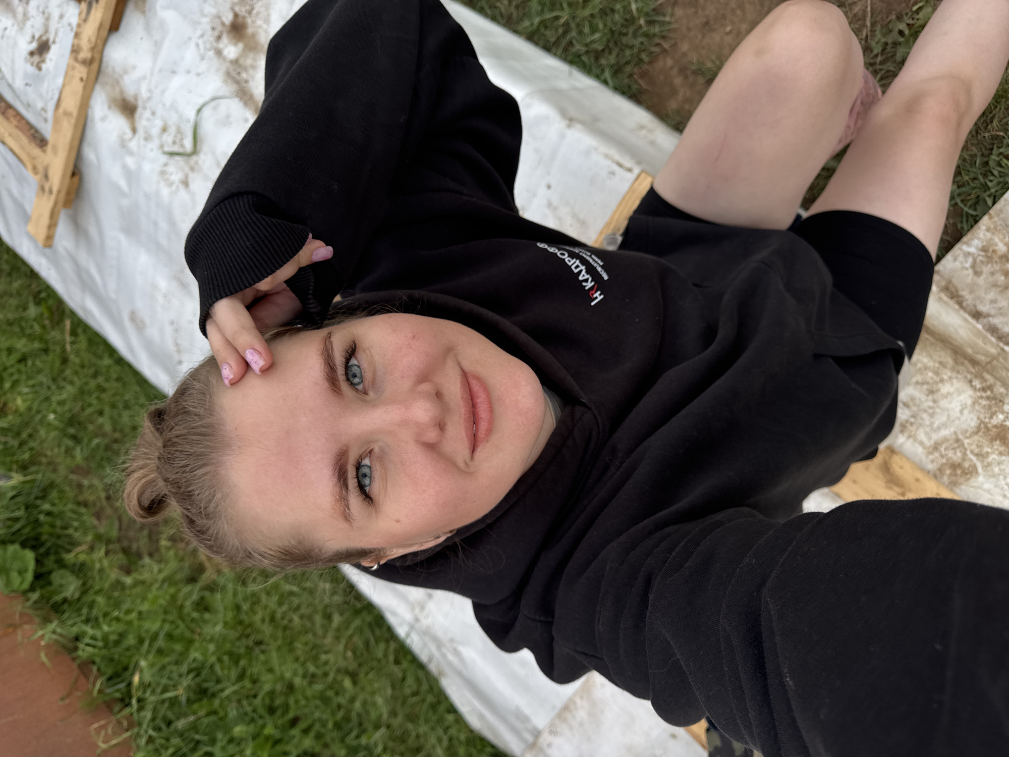 A young woman with blonde hair pulled into a bun is lying on her back outdoors. She is wearing a black hoodie and black shorts. Her eyes are blue and she is smiling at the camera. Her hand is resting on her forehead, and her fingernails are painted pink with a subtle design. The background consists of grass and a white tarp, suggesting an outdoor setting like a farm, possibly in Russia, given the text on her hoodie.
The text on the hoodie, partially visible, appears to be "hr KADPOФ". This could be a brand name or part of a logo. The lighting suggests it is daytime, and the presence of grass indicates an outdoor environment. The woman's relaxed pose and smile convey a sense of ease and contentment. The overall scene is casual and relaxed, possibly a moment of rest or a candid photograph taken during an outdoor activity on a farm.