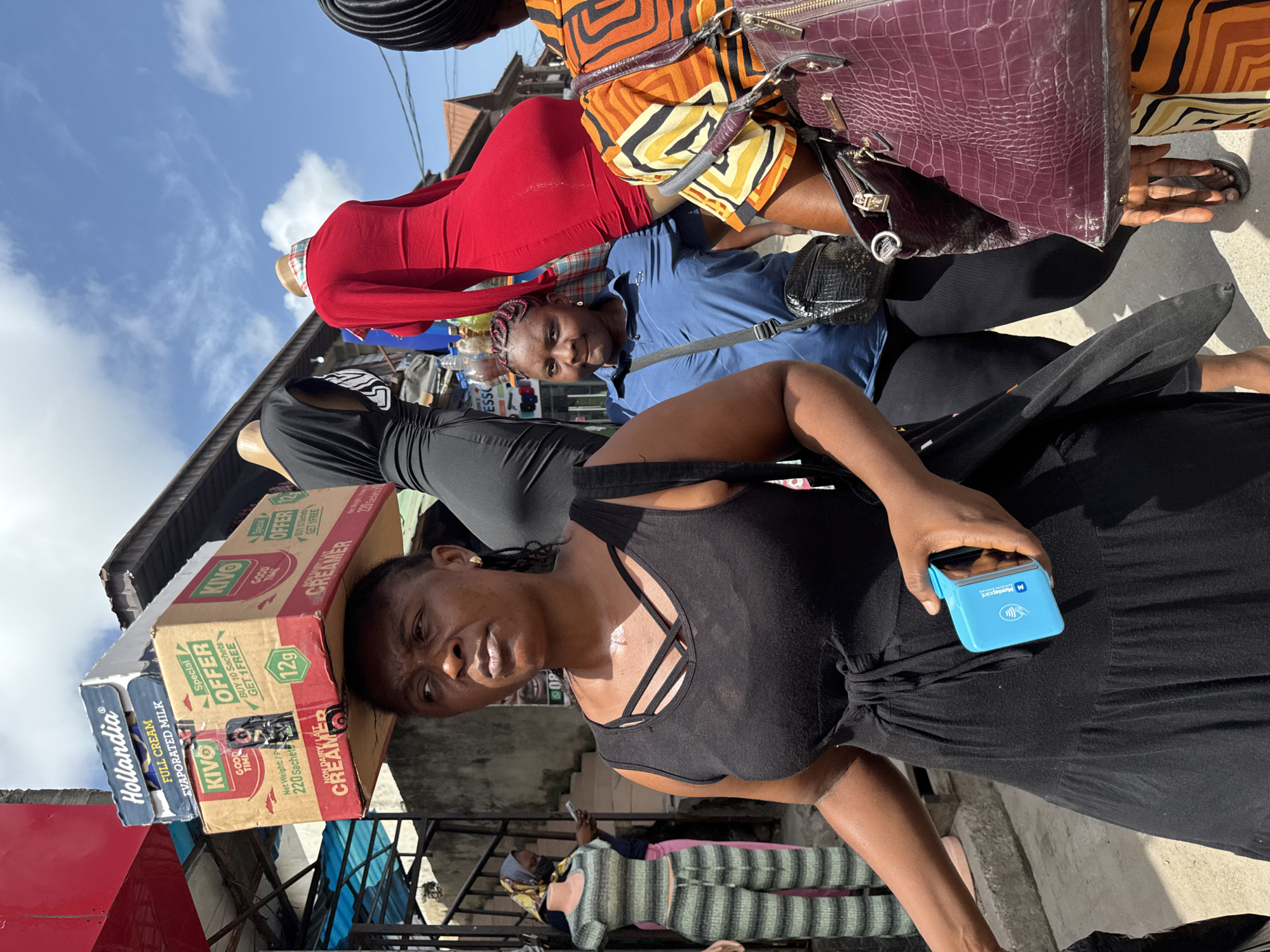 The image shows a woman in a black dress carrying boxes of Kivo Creamer and Hollandia Evaporated Milk on her head. She holds a blue Moniepoint device in her hand.
Behind her, several people are visible: a woman in a red dress, another in a blue shirt, and a third in an orange and yellow patterned dress. A woman in a striped top and headscarf is sitting on a bench behind a metal barrier.
The setting appears to be an outdoor market or commercial area in Ado, Nigeria. There are structures and various goods visible in the background.