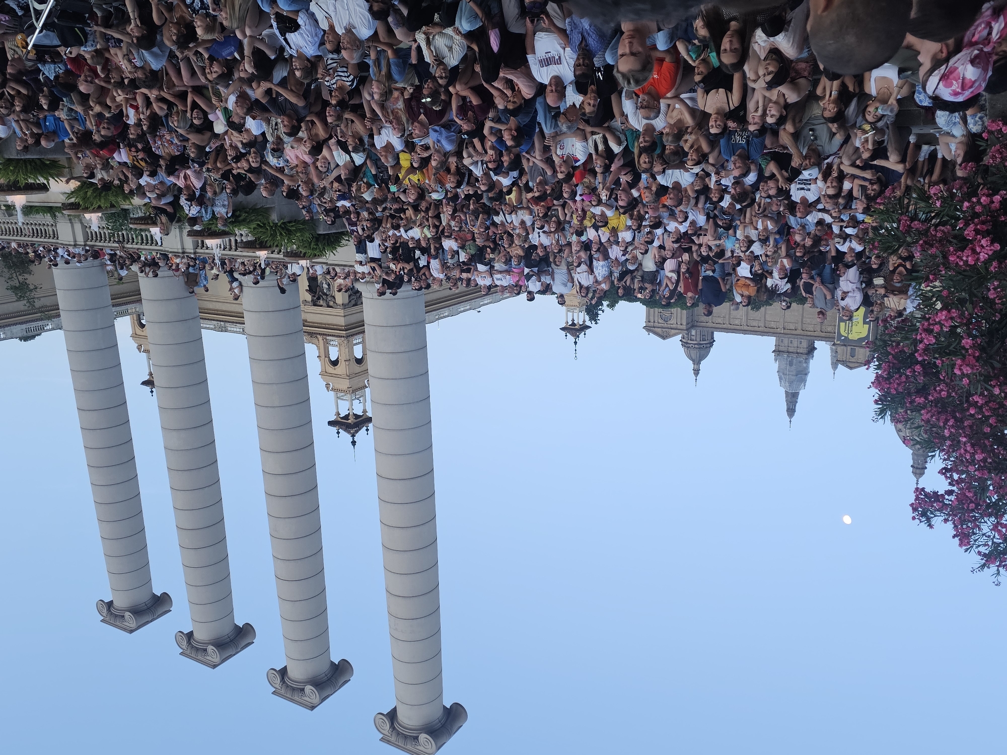 This image shows a large crowd of people gathered outdoors, likely for an event or performance, as they are all facing in the same direction. The scene is dominated by four large, fluted columns in the foreground, characteristic of classical architecture. In the background, glimpses of ornate buildings with domes can be seen, suggesting a significant historical or cultural location. A cluster of vibrant pink flowers is visible on the right side of the frame, adding a touch of natural color to the architectural setting. The sky is a clear, pale blue, and a faint moon is visible, indicating it might be late afternoon or early evening. The people are dressed in casual summer clothing, and their expressions are varied, some appearing engaged, others relaxed. The image is oriented vertically with the top of the scene appearing at the bottom. There is some text visible on a white t-shirt worn by a person in the crowd which reads "STAR". The overall impression is of a public gathering in a beautiful, historic urban setting.