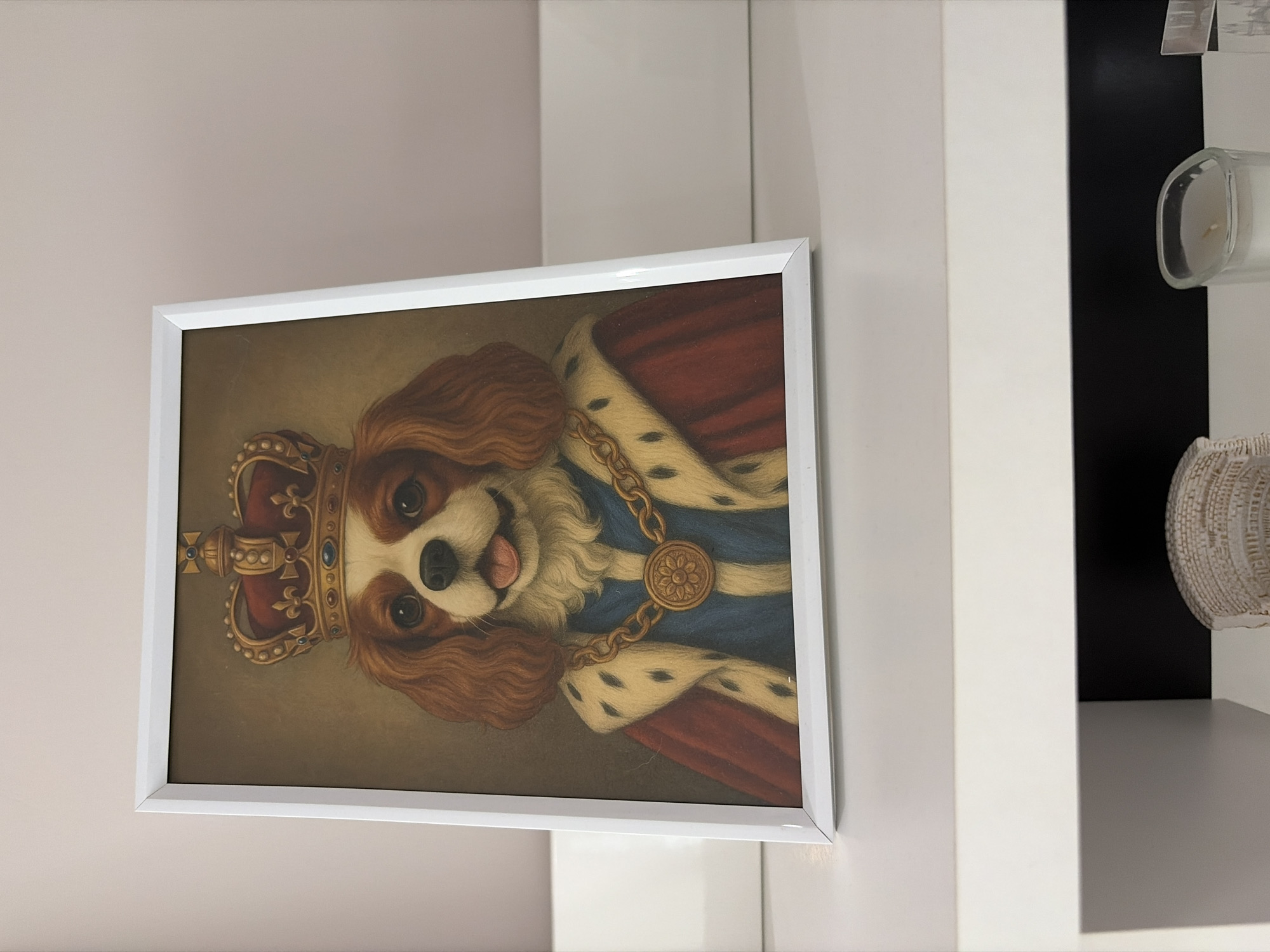 The image displays a framed portrait of a Cavalier King Charles Spaniel depicted as royalty, resting on a white shelf against a light pinkish-beige wall. The dog, with brown and white fur, large dark eyes, and a slightly visible pink tongue, wears a gold crown adorned with red velvet and blue/gold jewels. It is dressed in a blue garment, a red cape trimmed with white fur featuring black ermine spots, and a thick gold chain with a circular gold medallion displaying a floral design. The artwork style resembles a classical oil painting with a muted brown background. The frame is rectangular and white, leaning slightly back against the wall. Below the shelf, a lower white surface is partially visible, hosting a light-colored, textured woven object on the left and a clear, irregularly shaped glass candle holder with a white candle on the right. A dark recessed area is visible behind the objects on the lower shelf. The environment is an indoor setting with no specific geographical markers identifying a city or country, including Dobrich, Bulgaria.