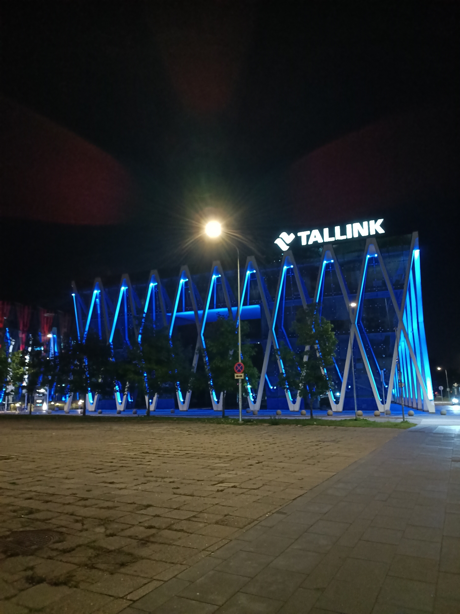 The image shows a modern building at night, illuminated with blue lights. The building has a distinctive geometric design with large glass panels and a prominent "TALLINK" logo at the top. Trees are visible in front of the building, and a paved plaza is in the foreground. The sky is dark, with a bright light, possibly a streetlamp or the moon, visible in the upper center of the image, creating a lens flare. The overall impression is one of a contemporary urban setting at night, likely a convention center or a prominent business establishment in Tallinn, Estonia.