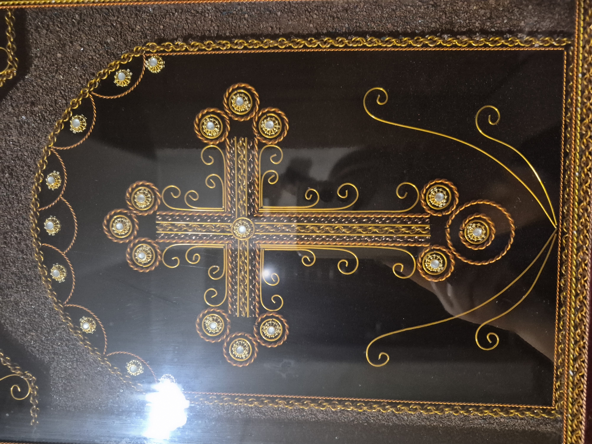 The image shows a close-up of a decorative metal cross. The cross is made of gold and copper wire, with small white jewels embedded in the center of each arm and at the intersection of the arms. The cross is set against a dark brown background, possibly velvet or a similar fabric, which is framed by a textured, granular material in shades of brown and grey. The entire piece appears to be a framed artwork. There is a reflection in the surface of the artwork, showing a person's hand and arm, indicating the image was taken by someone holding a camera or phone. The specific location in Vanadzor, Armenia, is not provided in the image or its context.